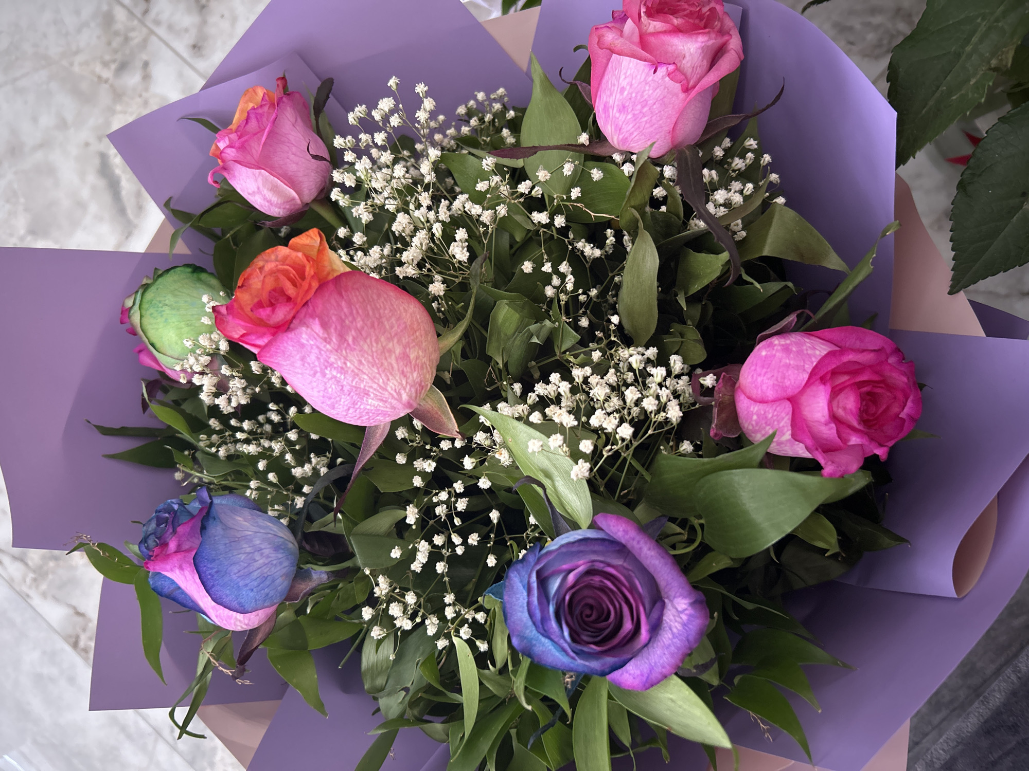 A close-up, overhead shot displays a bouquet of multi-colored roses and baby's breath, wrapped in purple paper. The roses feature an ombre effect, with colors ranging from bright pink and orange to blue and purple. One rose is predominantly green at the top. The bouquet is arranged in a shallow, circular formation, with the stems obscured by foliage and wrapping. The background is a muted, gray and white marbled surface. No people are present in the image. The location is the city of Pirşağı, Azerbaijan.