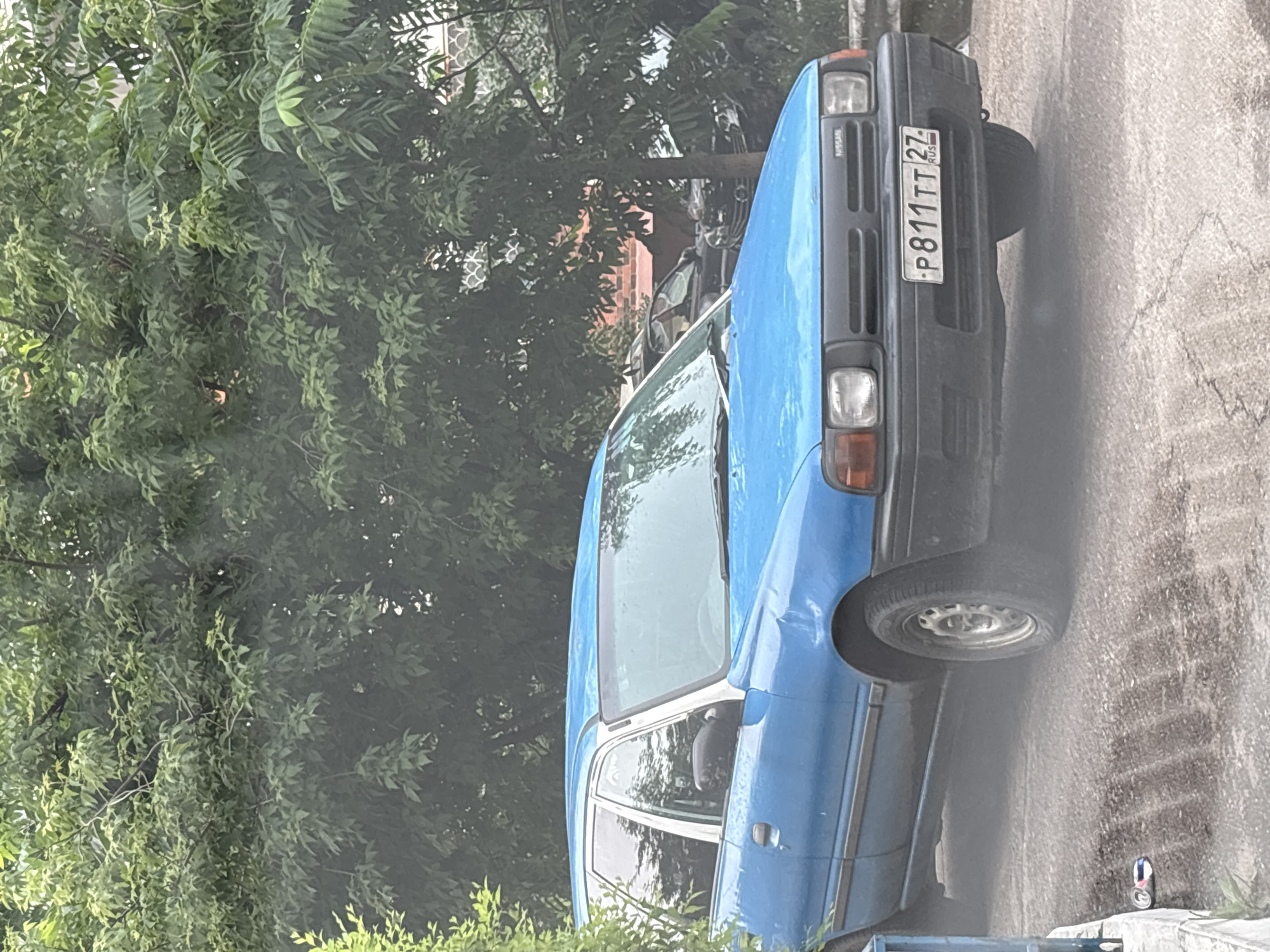 A blue Nissan station wagon is parked on an asphalt surface, partially obscured by a light haze. The car has a white roof trim and a black grille with "NISSAN" written on it. A license plate reading "P811TT 27 RUS" is attached to the front of the vehicle. Behind the car, dense green foliage from trees is visible, with hints of buildings in the background. The ground appears wet and somewhat dirty. An aluminum can is lying on the ground to the left of the car. The scene is consistent with an outdoor parking area in Blagoveshchensk, Russia, given the Russian license plate and general urban setting.