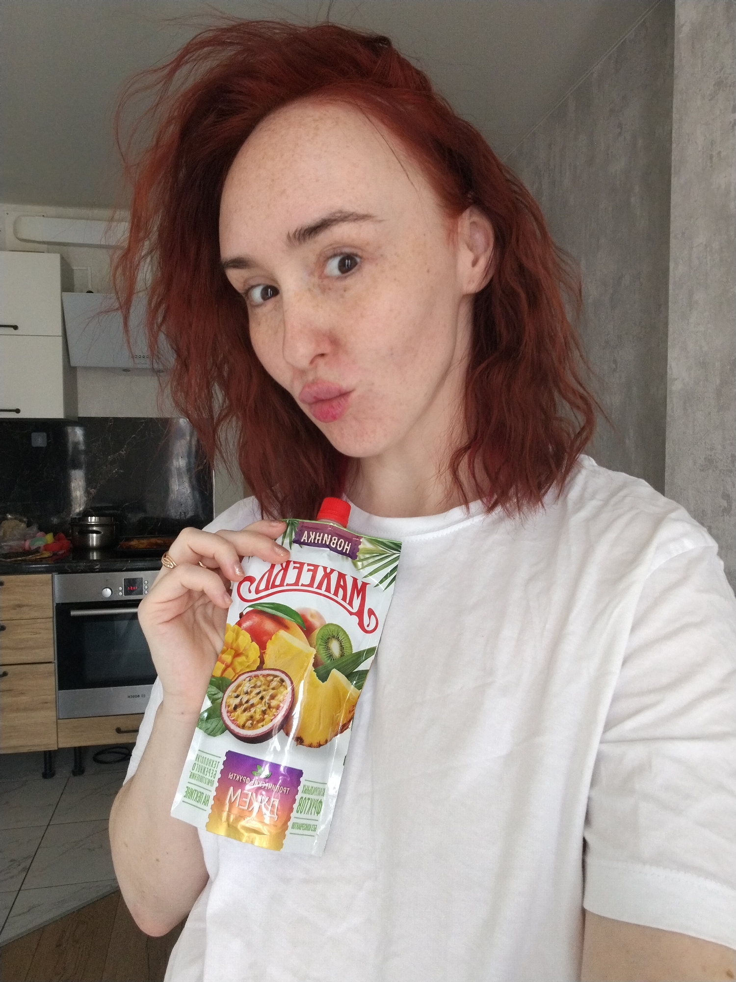 A woman with red hair and freckles is holding a package of "MAXLER" brand fruit juice in her left hand. She is wearing a white t-shirt and puckering her lips. The juice package has images of mango, passion fruit, kiwi, and pineapple on it. The text on the package is in Russian. She appears to be in a kitchen, with an oven and kitchen cabinets visible in the background. The wall behind her is a gray color. The location is Izhevsk, Russia, based on the product packaging.