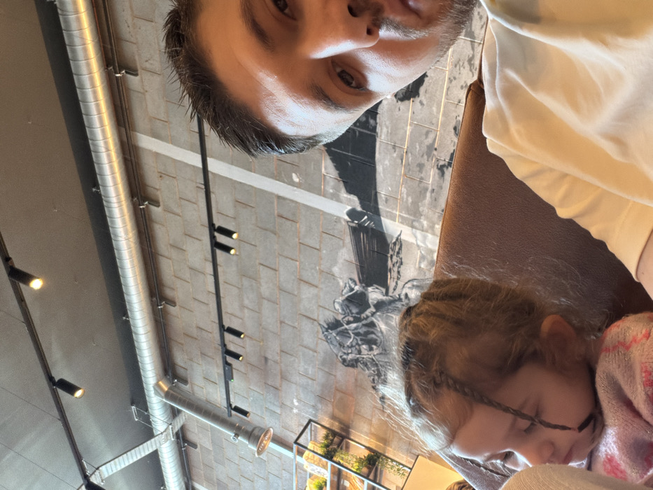 An adult male captures a close-up image of himself and a young child within an indoor establishment in Bretton, United Kingdom. The male, partially visible on the right with dark hair and a light-colored t-shirt, holds the camera, capturing his face from the eyes down, looking directly forward. To his left, a young child with light brown hair styled in multiple braids, some obscuring her face, appears to be sleeping or resting. She is wearing a pink, possibly knitted, garment. Both individuals are seated on a brown, upholstered booth or bench.
The background features an industrial-style interior. The ceiling is gray with exposed silver-colored metallic ductwork running horizontally and vertically. Multiple black track lights illuminate the space. The wall behind them is composed of light gray rectangular blocks, possibly concrete, and features a large, monochrome mural depicting a historical or maritime scene, including what appears to be a horse and parts of a boat. A black metal shelving unit with greenery is visible in the far left background.