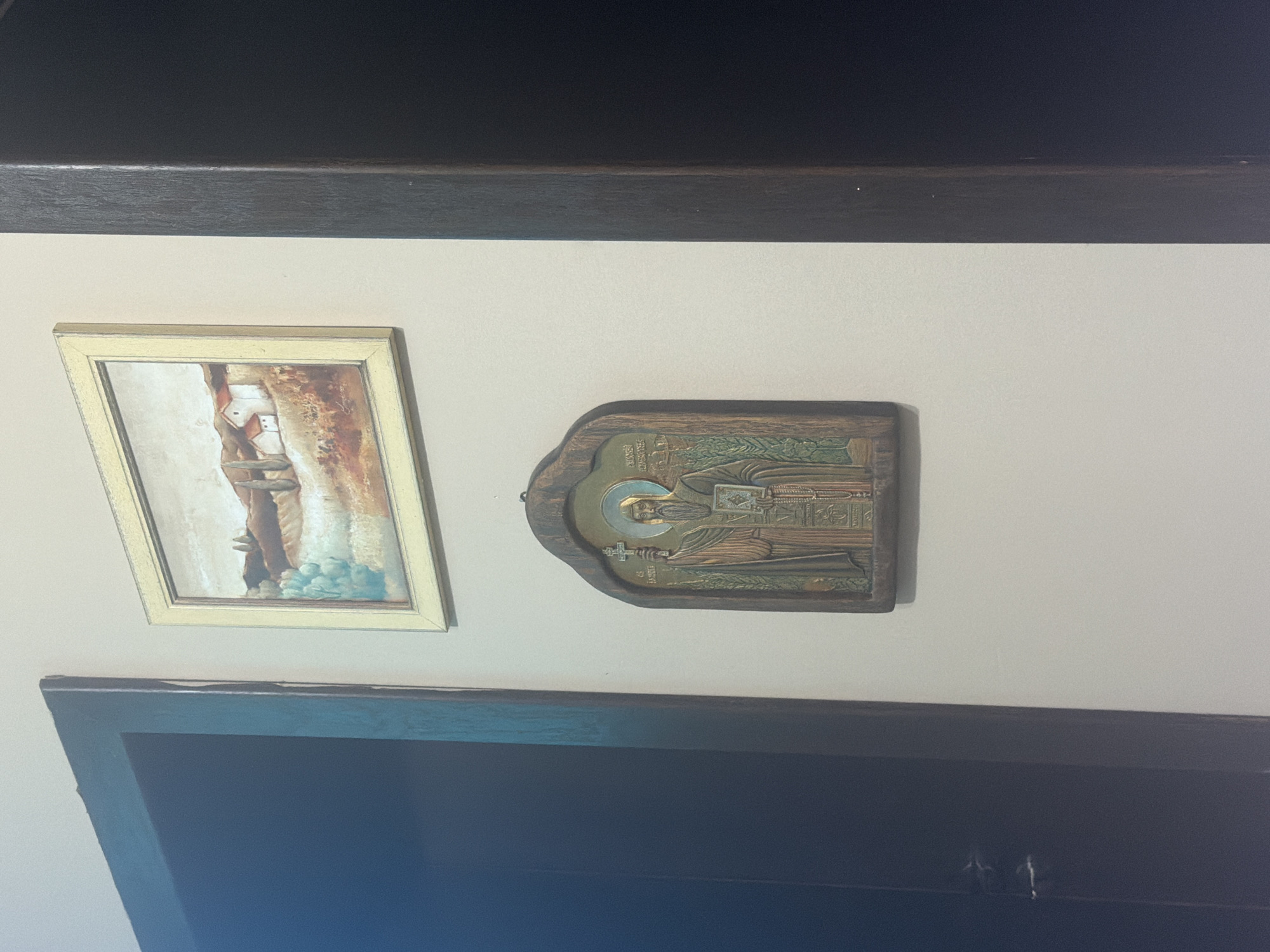 The image presents an interior wall, likely within a home or private space in Shumen, Bulgaria. Two framed artworks are displayed on a light-colored wall, situated beneath a dark wooden beam or shelf that extends across the top of the scene.
On the left, a rectangular picture, possibly an oil painting or print, is encased in a light-colored frame, perhaps cream or gold. The artwork itself features an abstract or impressionistic composition, dominated by vertical strokes and forms in earthy browns, whites, and touches of pale blue or turquoise. The specific subject is not clearly discernible but evokes a stylized landscape or figure.
To the right of this painting hangs a religious icon, characteristic of Orthodox Christian art. It is a wooden plaque with an irregular, arched top, featuring a relief sculpture or metal appliqué of a male saint. The saint is depicted with a prominent halo, a long beard, and monastic attire. He holds an open book in his left hand and a cross in his right. The icon incorporates metallic hues, primarily gold and bronze/copper, for the figure, halo, and background details. Cyrillic text is visible on the icon: "СВ. САВВА" (St. Savva) is clearly inscribed vertically to the left of the saint, identifying him as Saint Sabbas the Sanctified. Additional text, such as "СВЯТЬIЙ" (Saint) near the halo and "ОСВЕЩЕНИЙ" (Sanctified) on the right, further supports this identification.
Below the artworks, a dark, reflective surface, possibly a television screen or a piece of furniture, is partially visible, showing small bright reflections. The overall lighting suggests an indoor setting. The scene is static, showcasing a decorative and religious display.
