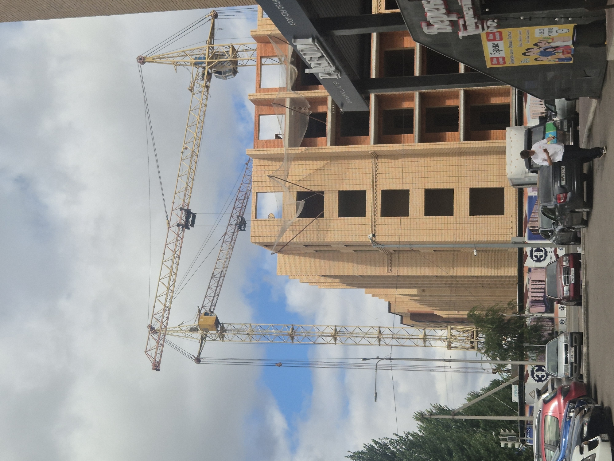The image shows a construction site in Kokshetau, Kazakhstan, featuring two yellow tower cranes against a cloudy sky and a partially constructed brick building. In the foreground, several cars are parked on a street. A young man in a white t-shirt and dark pants is walking near a truck, looking at his phone. A building with a prominent "PARFUM" sign is visible to the right of the construction site. The overall scene depicts an urban environment with ongoing development.