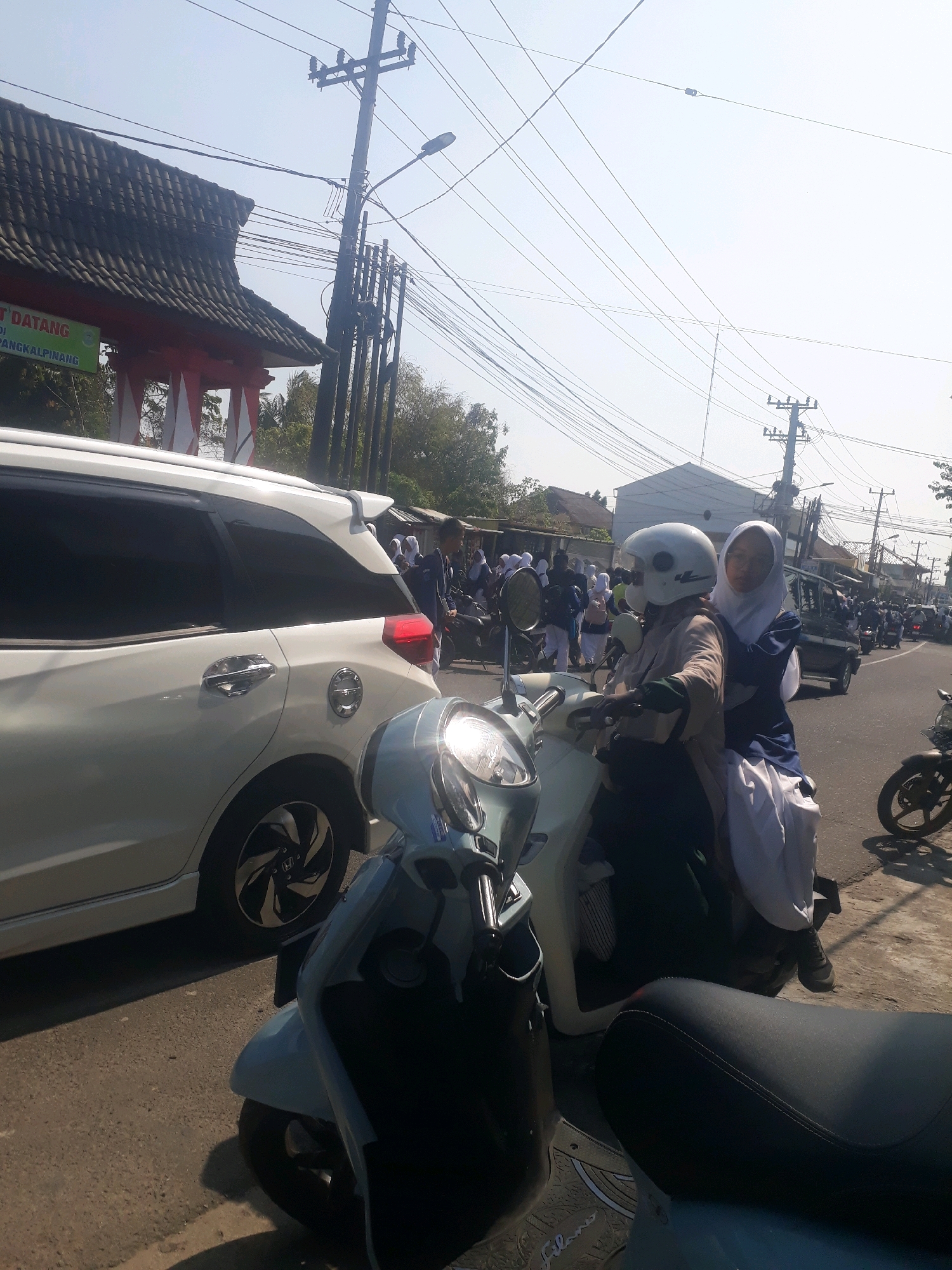 This image captures a scene on a sunny day in Pangkalpinang, Indonesia. In the foreground, two light blue scooters are parked, one closer to the viewer with its seat visible. Behind them, a white SUV is parked, its rear passenger-side door open. Further back, on the right side of the road, two young women are riding a scooter. The woman in front wears a light brown long-sleeved shirt and a white helmet, while the woman behind her wears a dark blue long-sleeved top and a white hijab. They are looking towards the viewer. A group of students, likely wearing school uniforms with white tops and dark bottoms or skirts, are walking on the sidewalk to the left, many carrying bags. In the background, there is a building with a dark tiled roof and red trim, possibly an entrance or gateway, with text that appears to read "SELAMAT DATANG DI PANGKALPINANG," indicating the location. Power lines crisscross the sky, and trees are visible. The road appears to be asphalt, and traffic is present with other motorcycles and a dark colored vehicle visible in the distance. The overall impression is of a busy street scene, possibly near a school or public gathering.