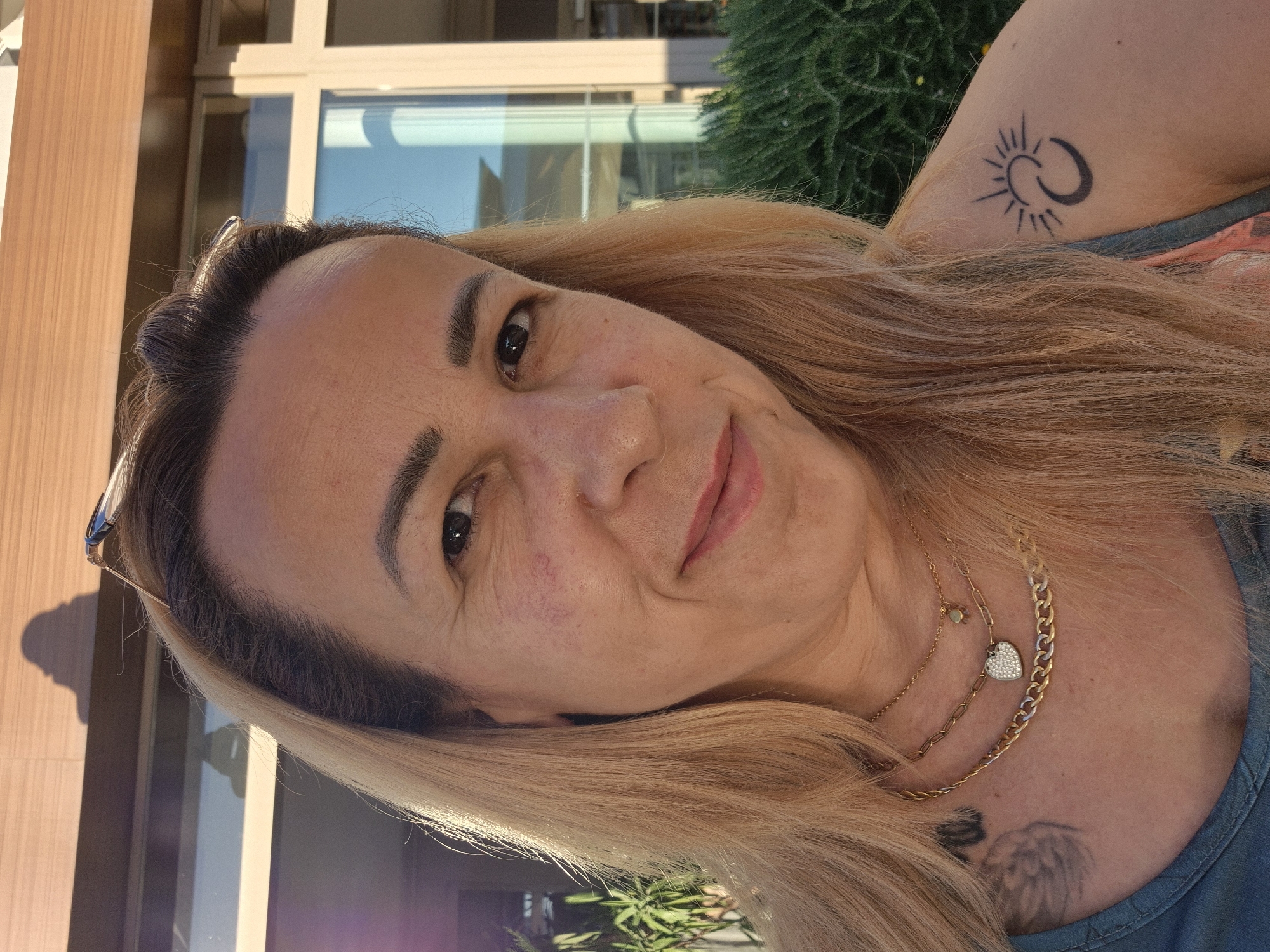 A medium shot shows a woman with blonde hair pulled back from her face, looking directly at the camera. She is smiling slightly, with her mouth closed. The woman is wearing multiple gold necklaces, one with a heart-shaped pendant and another with a chunky chain. She has a tattoo of a sun and moon on her left shoulder. Behind her, a building with a wooden facade and large windows is visible, along with some green bushes. The lighting suggests it is daytime, possibly late afternoon, with shadows cast by the building and a person, presumably the photographer, visible as a shadow on the building. The scene appears to be an outdoor setting, possibly a hotel or resort.