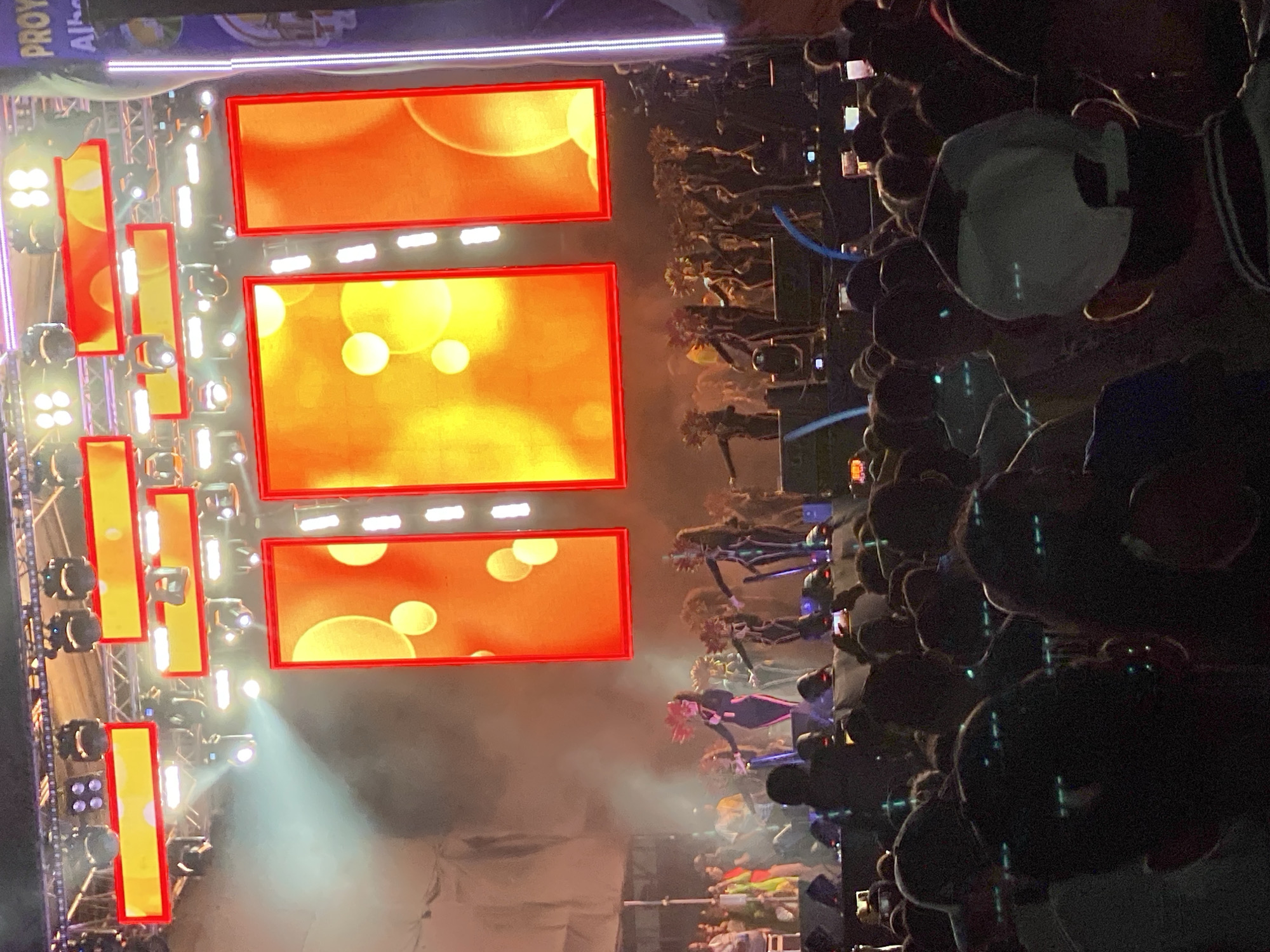 The image shows a large concert stage with three rectangular LED screens displaying abstract orange and yellow graphics. Vertical rows of rectangular lights flank the screens. Below the stage, a crowd of people is visible, some holding up phones. The stage is illuminated by bright spotlights. The event is located in Tinaquillo, Venezuela.