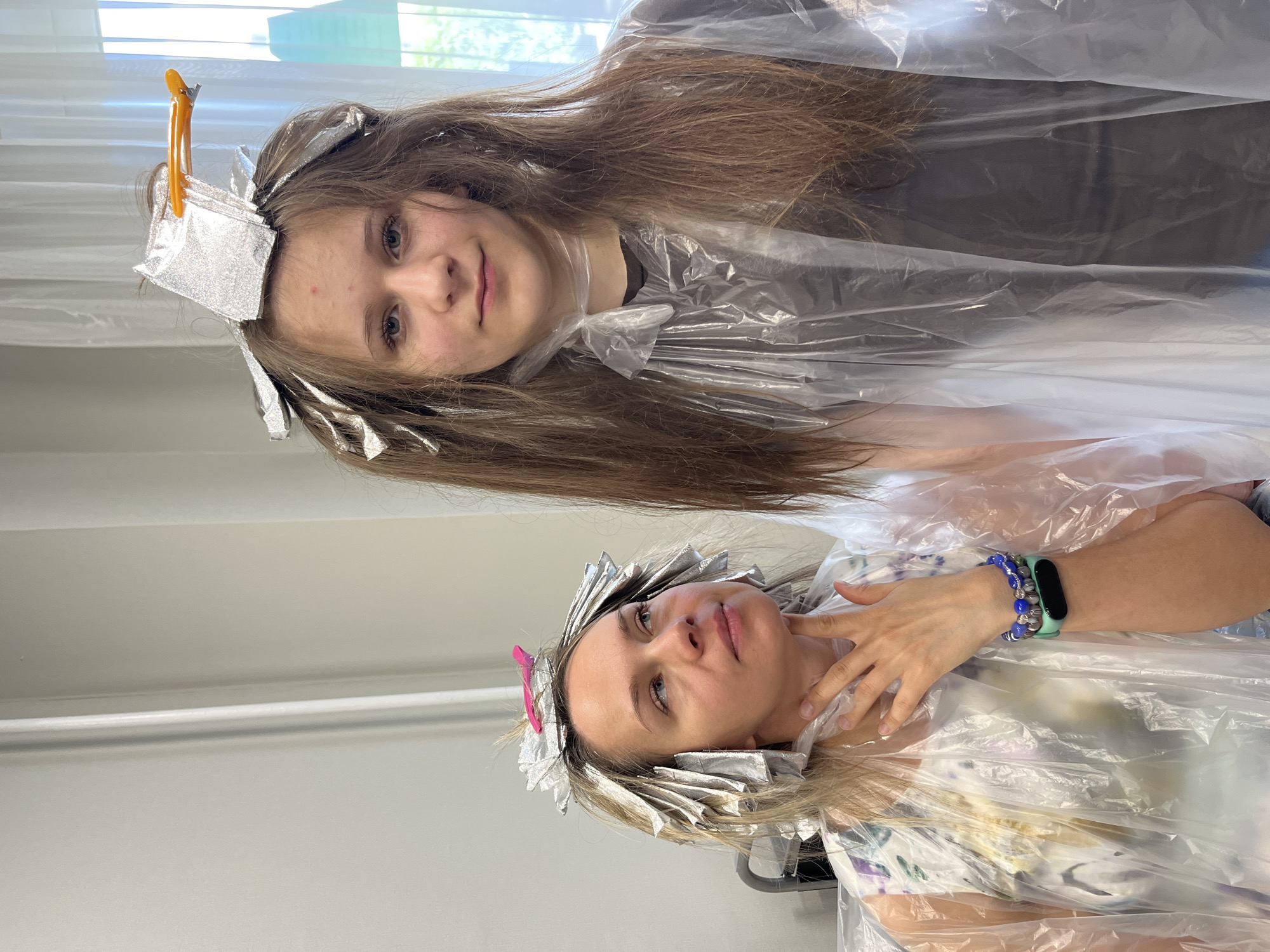 The image shows two individuals, likely a mother and daughter, in a room that appears to be a home or a salon. Both are wearing clear plastic capes, and sections of their hair are wrapped in aluminum foil with clips. The person on the right, a young female with long brown hair, is looking directly at the camera with a neutral expression. The person on the left, an adult female with blonde hair, is looking upwards and to the left, with her hand touching her chin. She is wearing a floral patterned dress. The room has light-colored walls and a white curtain in the background, suggesting an indoor setting. There is no visual information within the image to confirm a location in the city of Dubovoye, Russia.