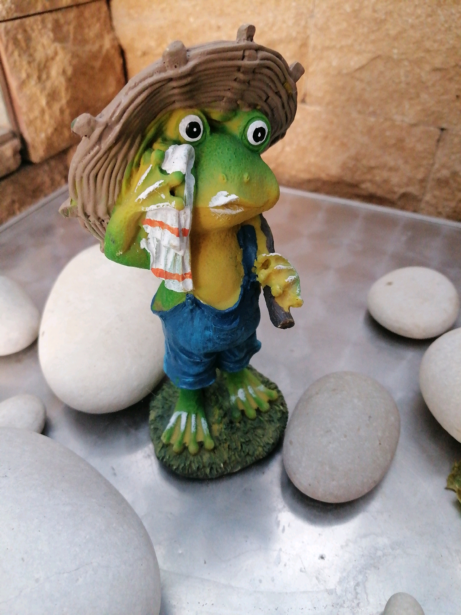 A ceramic figurine of a frog wearing overalls and a straw hat stands on a metal surface. The frog holds a white and orange striped cloth to its face, as if crying or wiping its eyes. It has large, round eyes with black pupils, and its skin is a vibrant green. The hat is textured and resembles woven straw. Several smooth, white stones are scattered around the figurine. In the background, a rough, textured stone wall is visible. The scene appears to be outdoors, possibly in a garden or patio area. There is no indication of its location within the city of Rostov-on-Don, Russia.