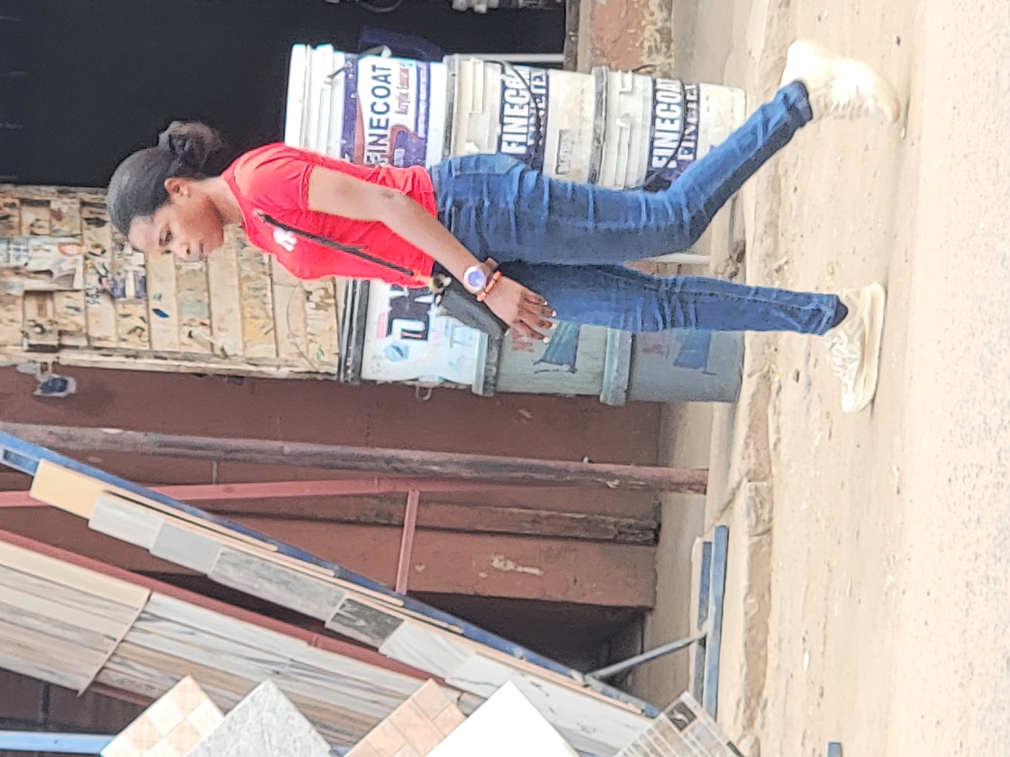 The image depicts a woman in Maiburiji, Nigeria, wearing a red top, blue jeans, and light-colored sneakers, standing in front of a building material store. The store has stacks of paint buckets with "FINECOAT" branding and displays of various tiles and construction materials. The woman has a watch on her left wrist and is looking down. The background includes the store's facade and a section of the street. The environment suggests a typical commercial area in Maiburiji.