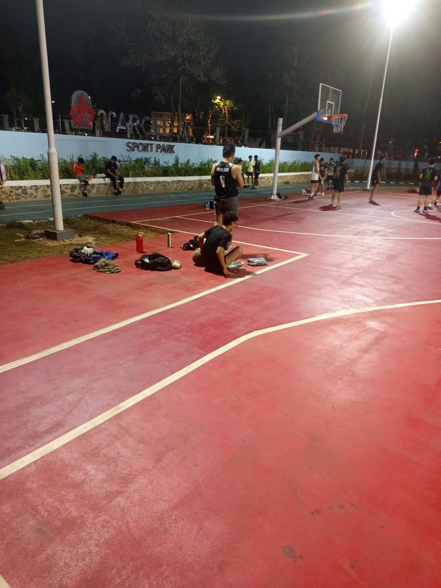 The image depicts an outdoor basketball court at night, illuminated by bright floodlights. The court surface is primarily red with white markings. In the background, a light blue wall features a large, metallic sign that reads "MARGO" in prominent letters, with "SPORT PARK" written below it. Several people are present on and around the court.
On the court, a group of young men, likely teenagers or in their early twenties, are engaged in or observing a basketball game. One individual in a black jersey with the number "8" on the back is standing near the basket. Another person in a similar black jersey is seated on the court in the foreground, facing away from the camera. Several other individuals are scattered around the court, some standing and others seated on the perimeter.
To the left of the court, a blue track runs parallel to a grassy area. On the grass, near the court's edge, several items are placed, including a red water bottle, a silver thermos, a black backpack, and some folded clothing. In the far background, trees are visible, their branches silhouetted against the night sky. The overall scene suggests a casual recreational activity taking place in a public sports facility. The presence of the "MARGO SPORT PARK" sign strongly suggests the location is in West Jakarta, Indonesia, as Margo City is a well-known shopping mall and entertainment complex located in Depok, which is adjacent to Jakarta, and "Margo Sport Park" is a known facility associated with it, often drawing visitors from the broader Jakarta metropolitan area including West Jakarta.