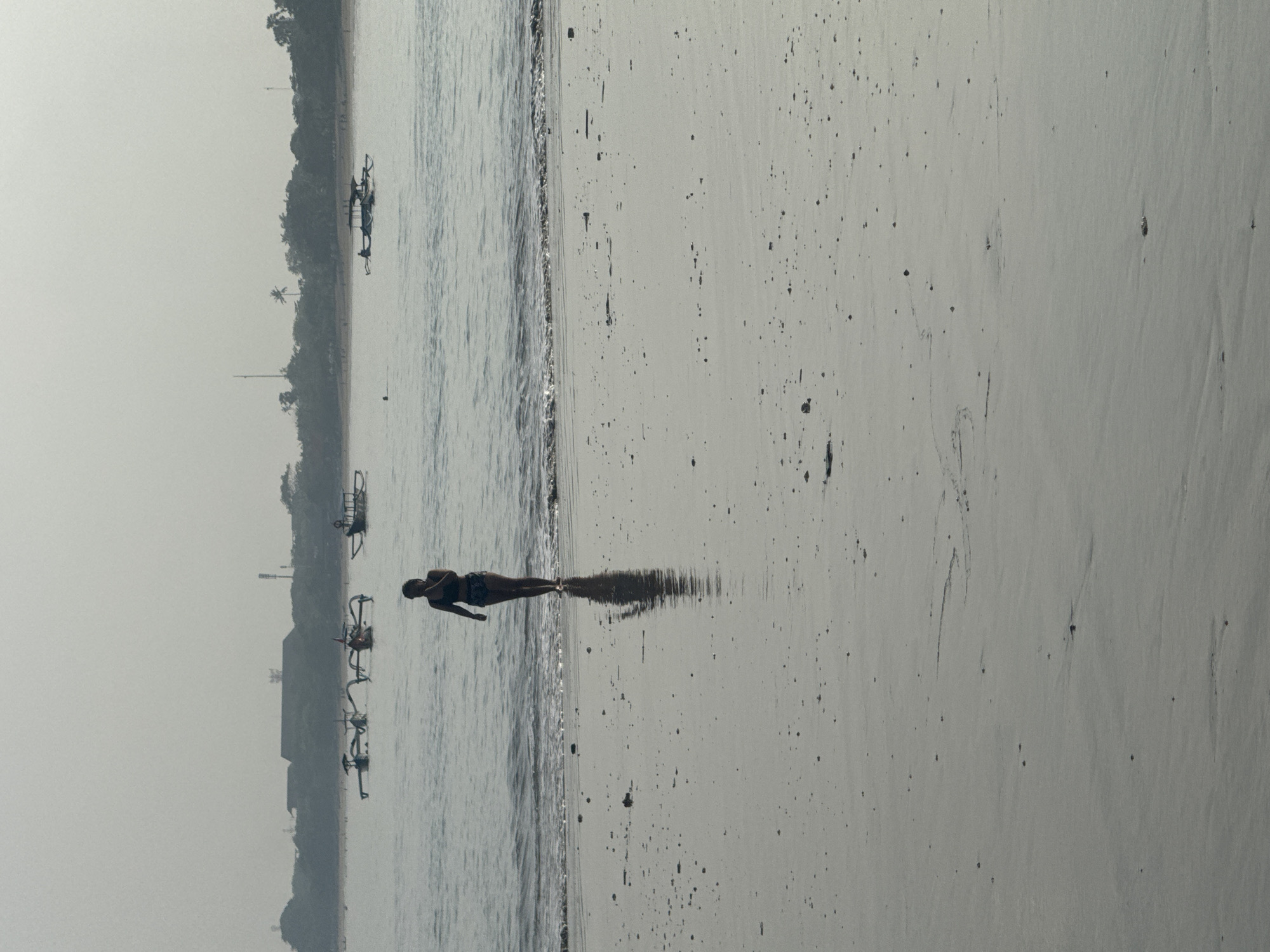 The scene depicts a beach landscape in Jimbaran, Indonesia. A body of water borders the shoreline, and a person is standing in the water near the edge of the beach. The person appears to be reflected in the wet sand. Along the coast, there are trees, buildings, and several small boats. The sky is overcast. The beach sand is damp, with scattered dark spots.