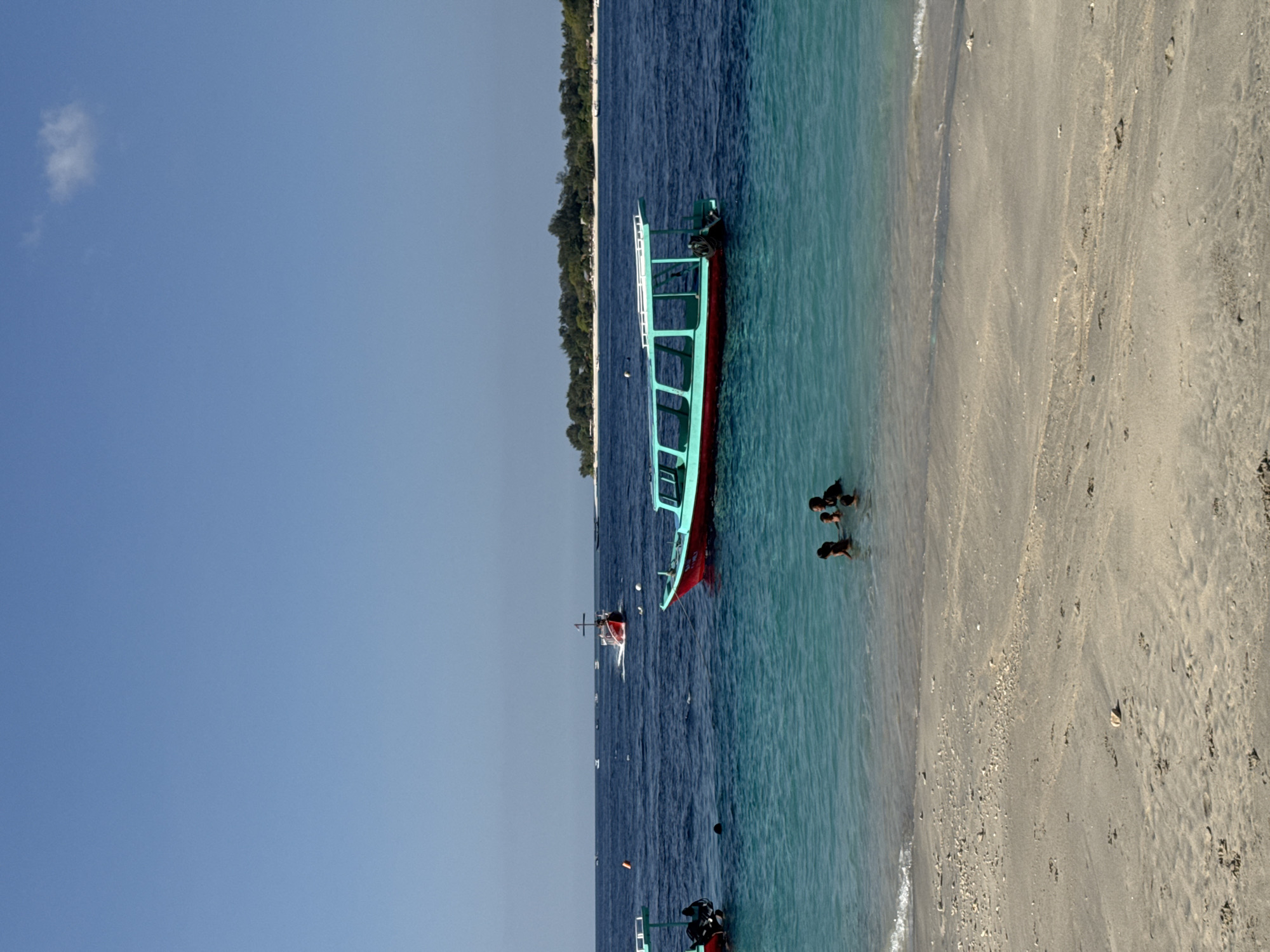 An eye-level, medium shot captures a tropical beach scene with clear blue skies and turquoise waters. A sandy shore stretches across the foreground, marked with scattered shells and footprints. The shallow water near the shore is transparent, revealing the sandy bottom. A group of four individuals, appearing to be children or young adults, are partially submerged in the water, their upper bodies visible as they interact.
Further out in the deeper blue ocean, two boats are present. A larger, turquoise and red boat with a covered seating area is moored or anchored. Another smaller, red boat with a white canopy is in motion, leaving a wake behind it. In the distance, a lush green island with a line of trees and some structures can be seen against the horizon. Several other smaller boats are scattered across the water, and a single orange buoy floats near the larger turquoise boat. The overall impression is a serene and picturesque day at a tropical destination.