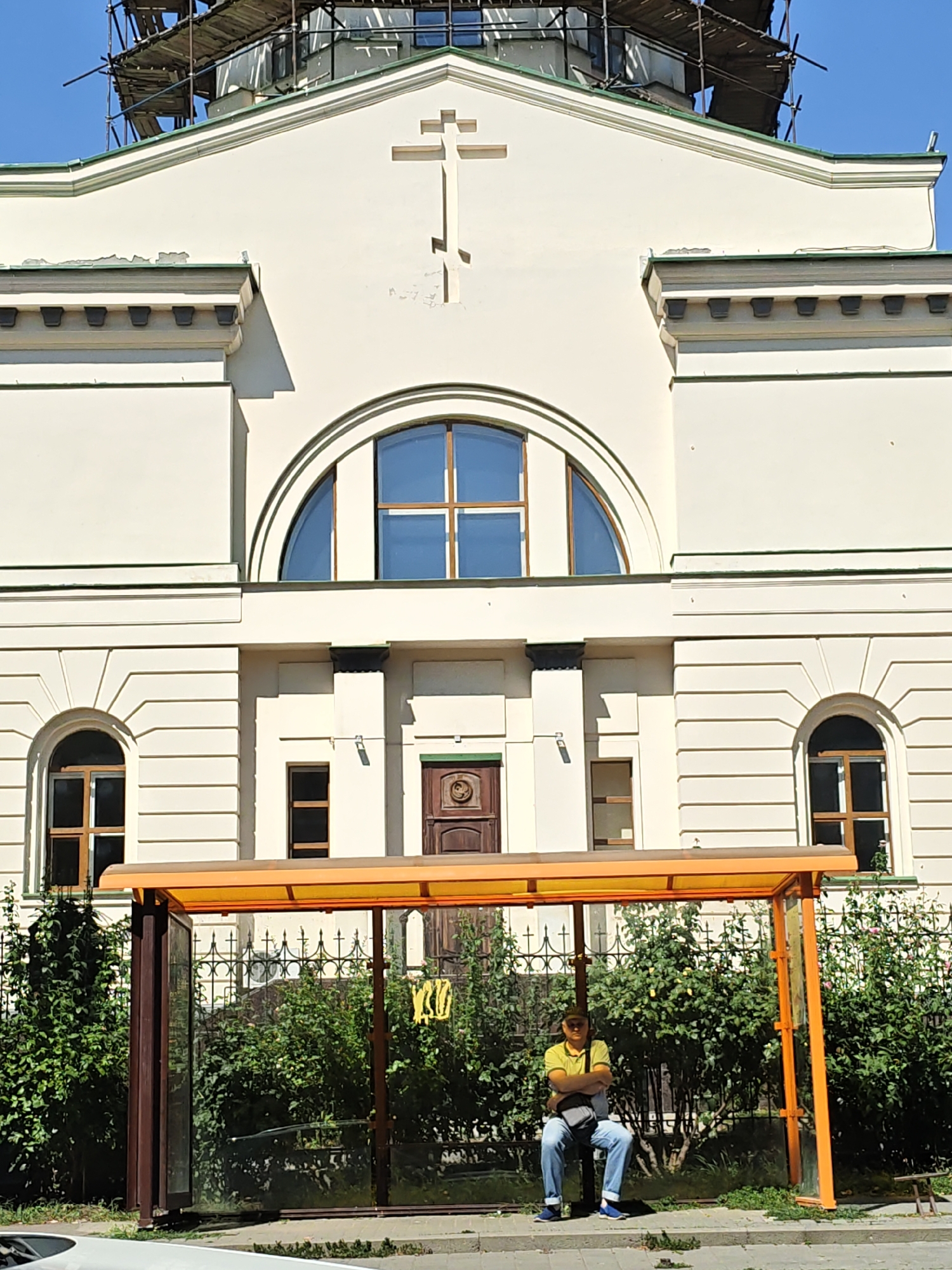A man sits at a bus stop in Rostov-on-Don, Russia, awaiting transportation in front of a large, pale-yellow building. The building appears to be a church, with a cross prominently displayed above a large arched window. The man is wearing a yellow polo shirt and blue jeans, and has a gray messenger bag slung across his body. He is sitting on a bench at the bus stop, which has an orange roof and glass walls. Behind the bus stop, a wrought-iron fence separates the bus stop from a lush green hedge. The building is under construction, with scaffolding visible on the roof. The sky is clear and blue.