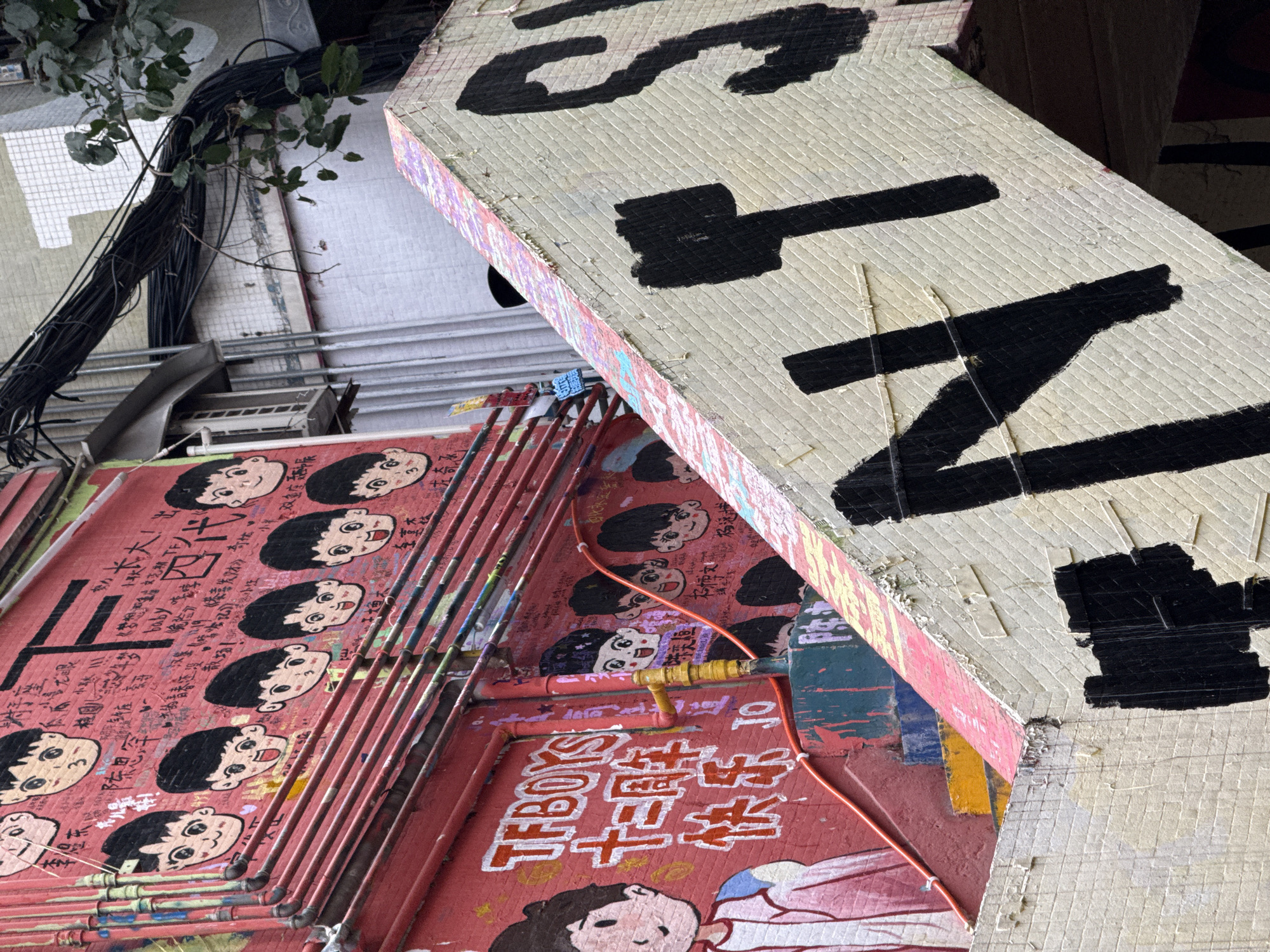The image depicts a densely urban scene in Jiulongpo District, China, characterized by vibrant street art and exposed utility infrastructure. A prominent red brick wall, heavily adorned with murals and graffiti, dominates the left and central portions. This wall features numerous cartoon-style heads of young boys with dark hair and expressive faces, some marked with the number "3" or "30" on their cheeks. Red, yellow, blue, and green conduit pipes run horizontally and vertically across this red wall. Large white text spelling "TFBOYS" and "十二周年快乐" (meaning "Happy Twelfth Anniversary") is visible on the lower left of the red wall, alongside a partial mural of a boy in white and pink clothing. A large black letter "F" is painted near the top right of the red wall.
Diagonally cutting across the lower right portion of the frame is a light-colored, tiled structure, likely a staircase or ramp, which features large, bold black letters, forming part of a word like "NTS." The top edge of this structure displays faded multi-colored paint, including pink and purple hues. Below this tiled structure, sections of painted steps in red, blue, yellow, and green are visible.
In the upper background, a dense bundle of thick black electrical cables is visible, running along a lighter-colored building facade, which appears to be tiled. A small cluster of green leaves from a plant branch is present in the upper right corner. An air conditioning unit is mounted on an adjacent building wall in the mid-left background. The scene exhibits layers of paint, graffiti, and exposed urban utilities.