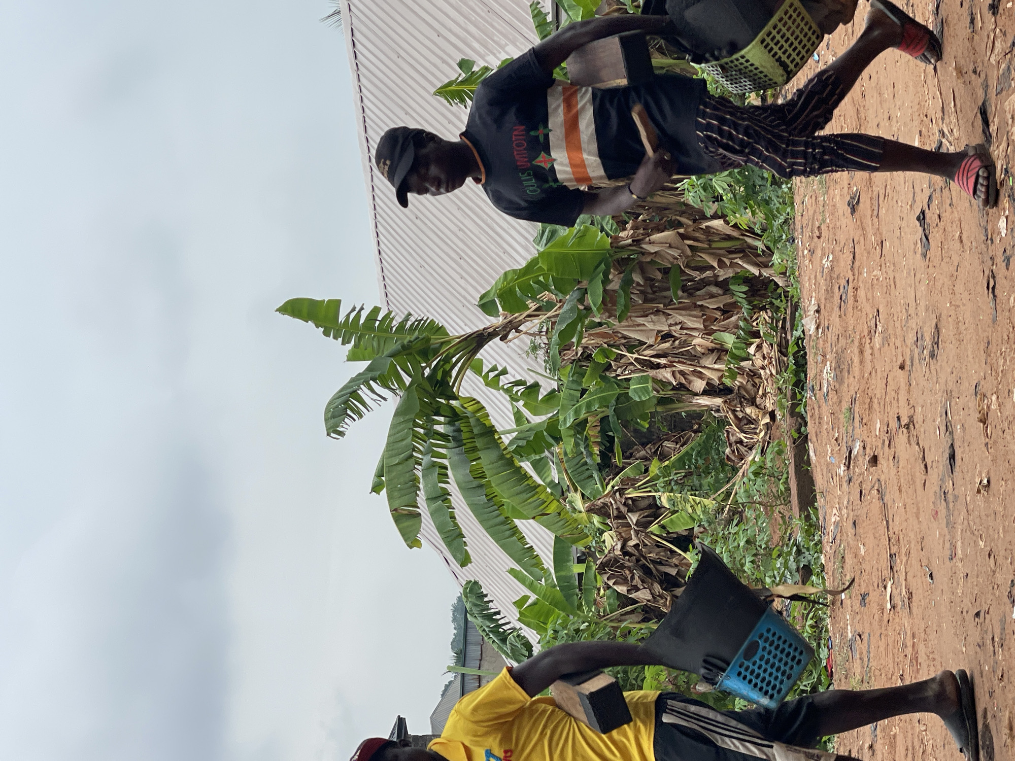 In Onitsha, Nigeria, two men carry baskets, presumably collecting items. The man in the foreground wears a yellow shirt, while the other wears a black shirt with orange and white stripes. Both wear sandals and dark-colored caps. They stand on a dirt path, surrounded by banana trees. In the background, a corrugated metal roof is visible under a cloudy sky.