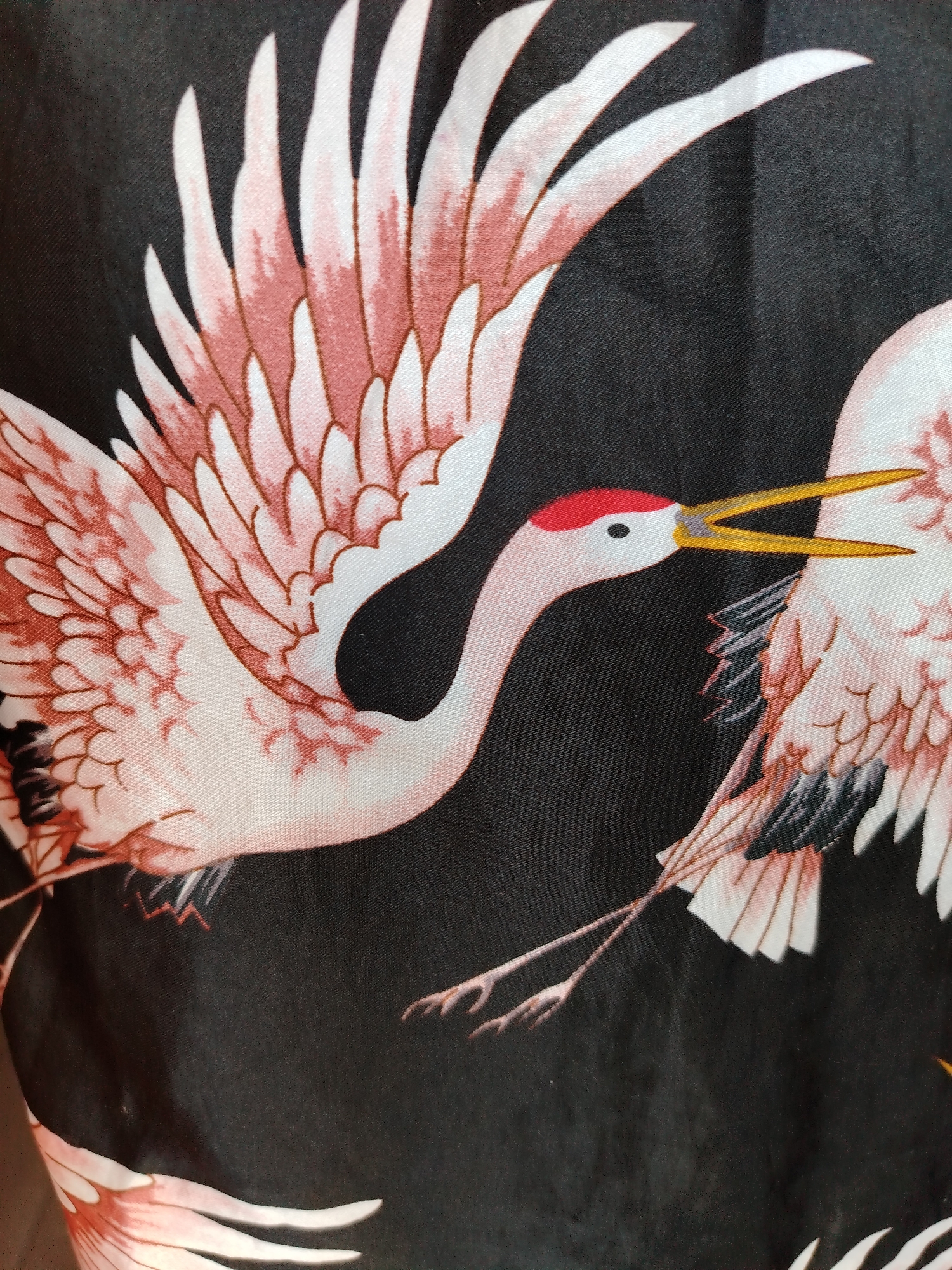 The image is a close-up of a fabric with a pattern of pink and white cranes on a black background. The cranes have red markings on their heads and long yellow beaks. Their wings are spread open. The image shows part of two cranes, with one crane's head and neck fully visible, while the other's is only partially visible. The fabric appears to be folded or draped. The image does not show a location in Izhevsk, Russia.