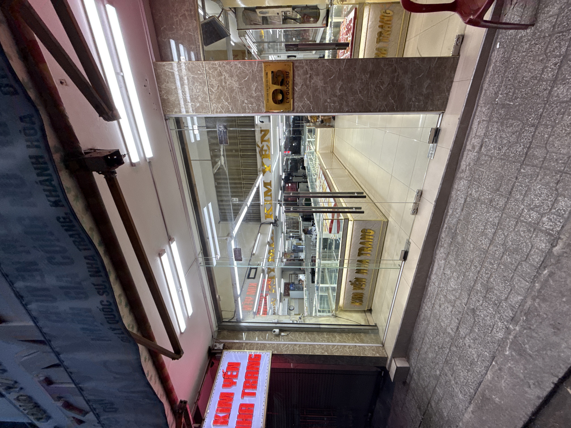 The image shows a storefront in Nha Trang, Vietnam, with the name "KIM YẾN NHA TRANG" displayed prominently. The storefront features a glass entrance with a golden sign that reads "05 LÝ QUỐC SƯ NHA TRANG." Inside, display cases filled with jewelry are visible, and fluorescent lights illuminate the store. A red chair is placed outside the entrance. Above the storefront, a banner with Vietnamese text is visible, partially obscured by the structure. The street in front of the store is paved with stone tiles.
