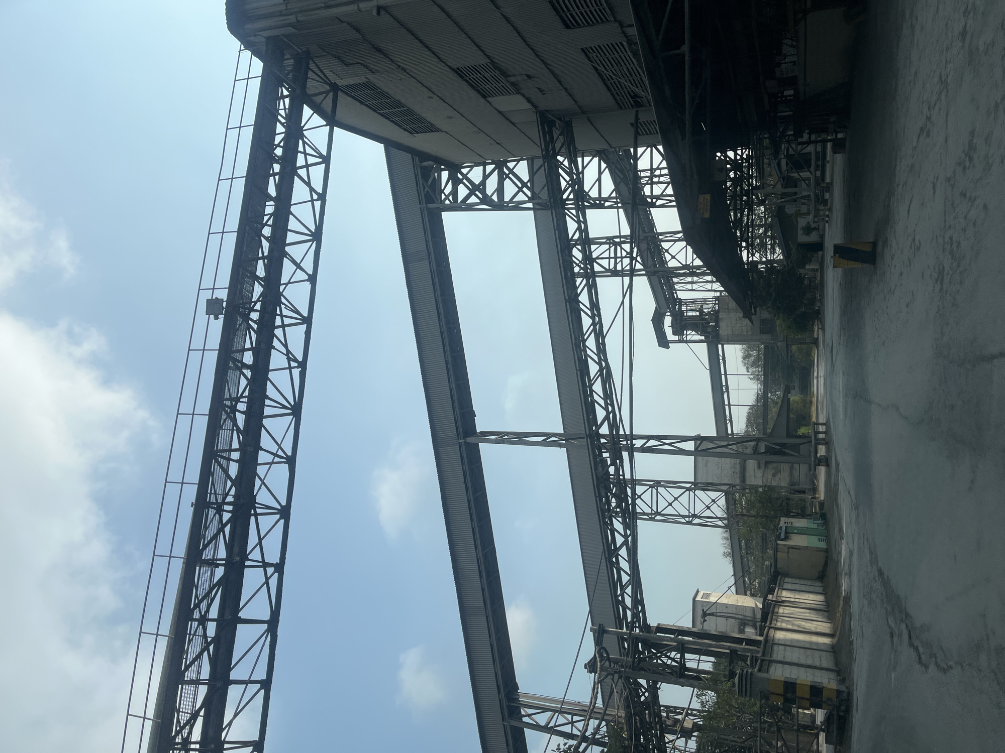 The image shows an industrial facility, likely a factory or processing plant, with a clear sky and some scattered clouds. Dominating the scene are multiple elevated conveyor belt systems, supported by a complex network of steel trusses and towers. The conveyors are covered, suggesting they are used to transport materials. On the right side of the image, a large building with corrugated metal siding and small, horizontally-slatted windows is visible. The ground in the foreground is paved and appears dusty or covered in fine particles, consistent with an industrial environment. In the background, more structures of the facility can be seen, along with some trees. There are no people or animals visible in the image. The overall impression is one of a large-scale, operational industrial complex.