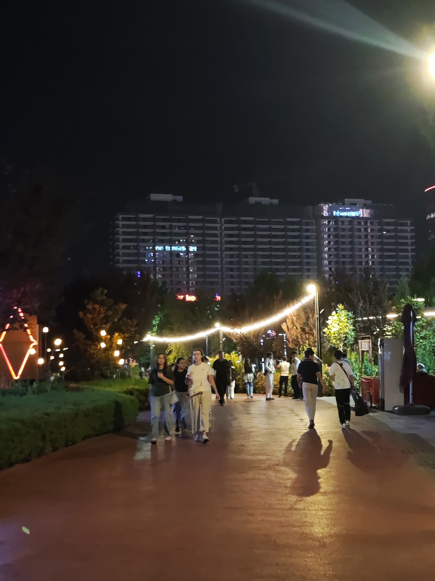 This image captures a nighttime scene in Tashkent, Uzbekistan, likely in a modern urban park or pedestrian zone. In the background, two large, multi-story buildings dominate the skyline, illuminated by countless windows, suggesting a commercial or residential complex. The buildings appear to have some neon or LED signage on their facades. In the foreground, a paved pathway, possibly a walkway or promenade, is visible, wet and reflecting the ambient light. A group of people, appearing to be a mix of locals and tourists, are walking along this path. They are dressed in casual attire, with some carrying bags. The pathway is bordered by lush green bushes and trees, suggesting a landscaped area. Decorative lighting, in the form of string lights and illuminated decorative elements, is present, enhancing the nighttime ambiance. The overall impression is one of a lively, well-lit urban space at night.
