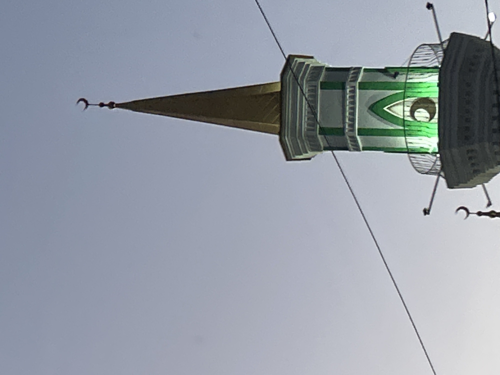 The image captures a minaret of a mosque in Orsk, Russia, against a clear, pale sky. The minaret is tall and slender, tapering to a golden spire. It is decorated with green and white accents, and illuminated by green lights. A crescent moon emblem is visible on the spire and at the base of the illuminated section. A metal railing encircles a balcony partway up the minaret. Several thin wires are strung across the frame, obscuring parts of the minaret. The lower portion of the minaret is visible, showing a more intricate architectural design with a gray, octagonal base. The overall scene is still and devoid of any people or activity.