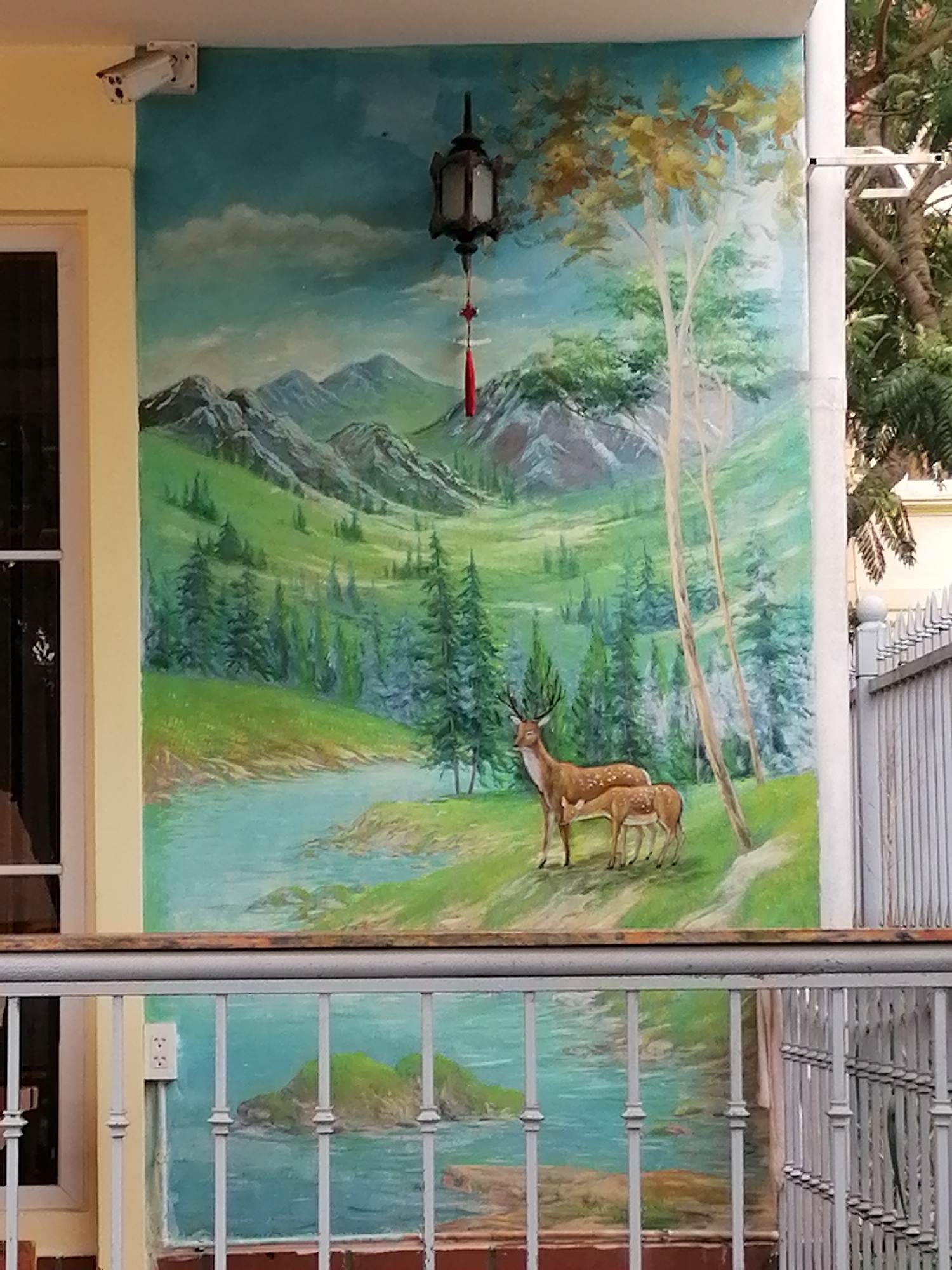 A vibrant mural depicting a serene mountain landscape adorns an exterior wall, likely in Cam Ly - Đà Lạt, Vietnam. The upper portion of the mural features a light blue sky with soft white clouds above a range of rugged mountains, rendered in blues and greens. Below the peaks, a lush green valley unfurls, dotted with numerous evergreen trees and a winding river or stream. In the foreground of this painted scene, a buck with antlers stands alongside a smaller fawn on a grassy bank beside the water, creating a tranquil pastoral image. Two tall, slender deciduous trees rise behind the deer on the right.
Above the mural, a white security camera is mounted on the top left corner of the wall. Centered above the painted landscape, an ornate, dark metal lantern with a red tassel hangs down. A light yellow architectural trim runs vertically along the left side of the mural, bordering a dark window. Below a metal railing that spans horizontally across the lower part of the image, the mural continues, showing a body of blue water with small green islands. An electrical outlet is visible on the yellow trim below the railing. To the far right, parts of a white pillar and a metal fence or gate, along with natural tree foliage, are visible, providing a contrast to the painted environment. The scene captures a peaceful daytime atmosphere.