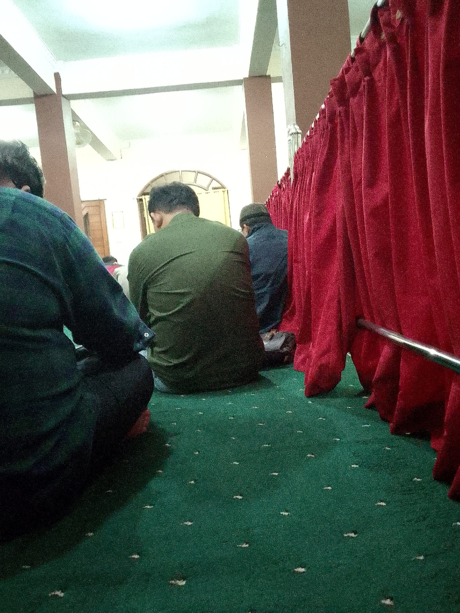 The image shows the interior of a mosque, likely in Kertajaya, Indonesia, during prayer. Three men are visible, facing away from the camera, seated on a green carpet with a speckled pattern. The man on the left wears a dark green long-sleeved garment, the man in the center wears a lighter green garment, and the man on the right wears a dark blue garment and a dark cap. A long, red curtain hangs on the right side of the frame, obscuring the background behind it. The lighting appears to be artificial, casting a diffuse glow from overhead fixtures. The overall atmosphere suggests a moment of quiet devotion within the mosque.