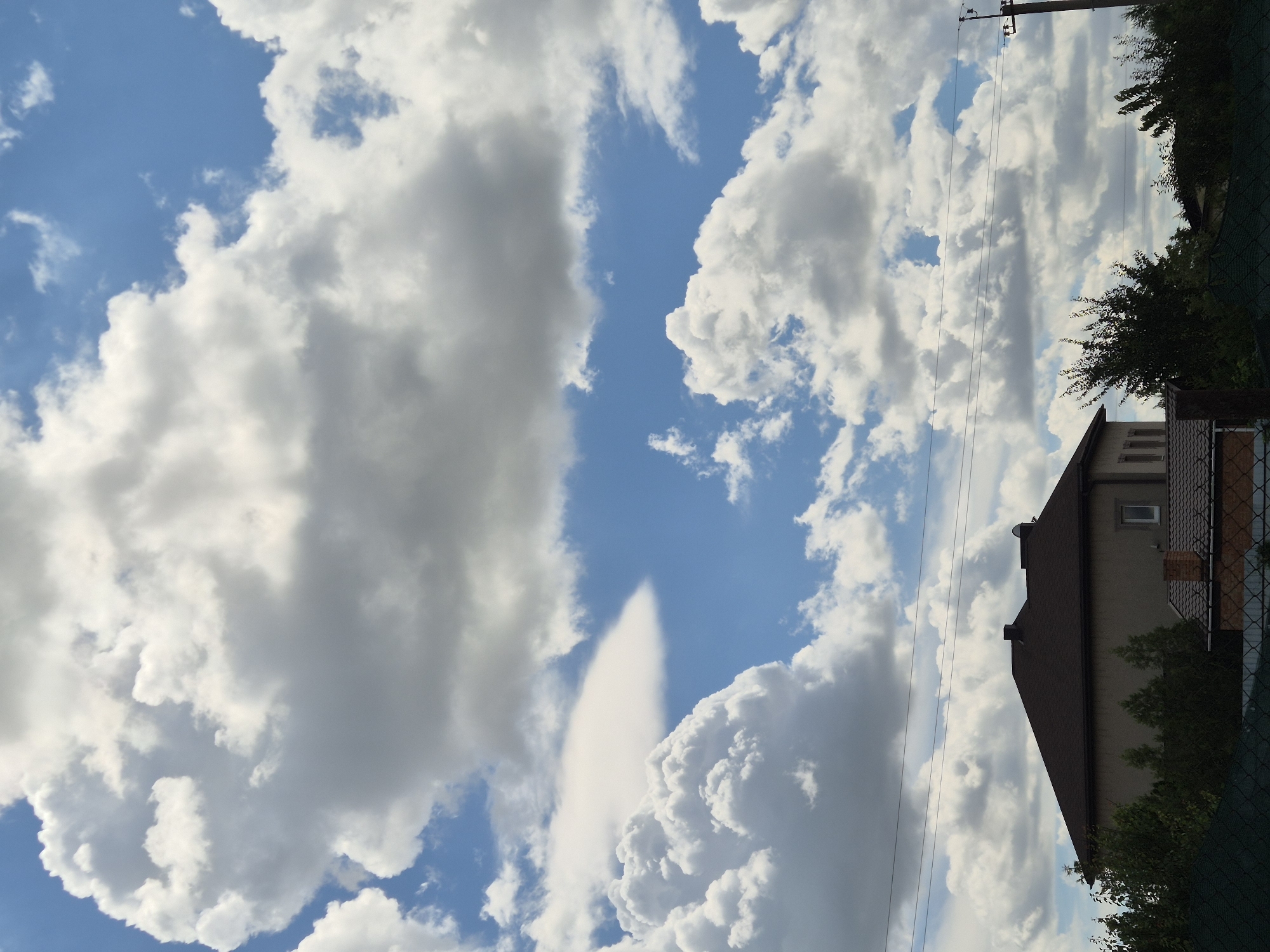The image depicts a daytime scene in Sozonivka, Ukraine, under a dynamic sky filled with large, fluffy white and grey cumulus clouds against a bright blue backdrop. In the foreground, a dark-roofed, two-story building with a light-colored facade and a small rectangular window is visible. Several evergreen trees and other foliage partially obscure the lower portion of the building. In front of this building, a smaller structure with a tiled roof and a brick chimney can be seen. A dark green fence runs along the bottom of the image. Overhead, multiple power lines traverse the scene horizontally. The overall impression is of a residential area on a cloudy, possibly summer day.