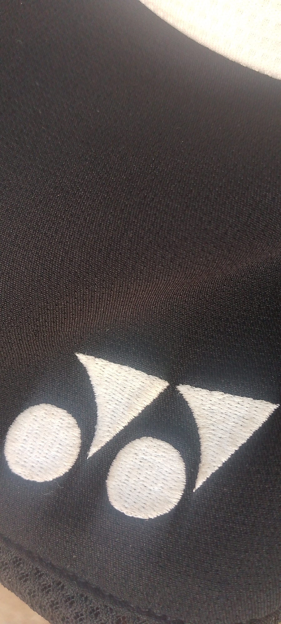 The image shows a close-up of a black garment with a white embroidered logo. The logo consists of three geometric shapes: two circles and two triangles arranged in a pattern that resembles an abstract representation of a stylized character or symbol. The embroidery is detailed and appears to be of good quality, with a visible texture from the stitching. The fabric of the garment is dark and appears to be a synthetic material, possibly polyester or nylon, with a subtle ribbed texture. The lighting in the image is somewhat diffuse, suggesting it might have been taken indoors or on an overcast day. There is no visible text in the image. The image is cropped in a way that focuses entirely on the logo and a portion of the garment, offering no information about the surrounding environment or any people. The setting is indistinct, but the overall impression is of a close-up detail shot of apparel.