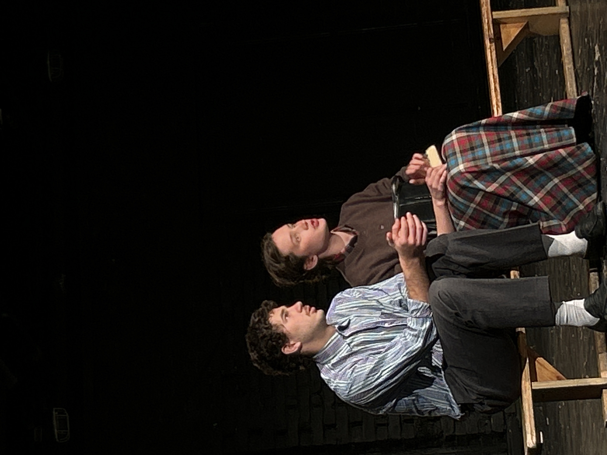 Two young individuals are seated on a wooden bench on a dimly lit stage. The person on the left has curly, dark hair and is wearing a striped button-down shirt and dark trousers. Their hands are clasped in their lap. The person on the right has medium-length, light brown hair and is wearing a brown sweater and a long, plaid skirt. They are holding a black rectangular object in their hands, which rests on their lap and appears to have a light-colored item on top of it. The background is dark and appears to be a brick wall, suggesting an indoor setting, possibly a theater or performance space. The lighting is focused on the subjects, creating a dramatic atmosphere.