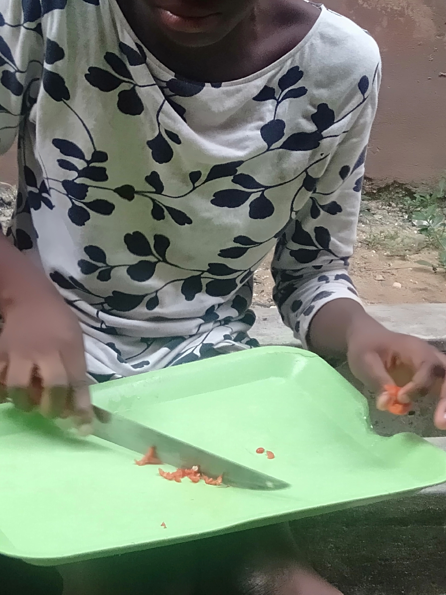 The image provides a close-up view of a person engaged in food preparation in an outdoor or semi-outdoor setting. The individual, visible from the chest down, has a dark skin tone and is wearing a long-sleeved, light-colored shirt featuring a distinct pattern of dark blue or black leaves and stems. Only the lower part of their face, specifically the mouth and chin area, is visible at the very top of the frame.
The primary activity depicted is the chopping of food items. The person is using a kitchen knife with a metallic blade to dice small, red-orange pieces of what appear to be chili peppers or possibly tomatoes. These pieces are being prepared on a light green, somewhat rectangular plastic tray or cutting board. The person's left hand is positioned near the knife and the chopped items, while their right hand holds another small, unchopped piece of the red-orange item, ready to be added to the tray. Numerous small, irregularly shaped chopped pieces are scattered on the green surface of the tray.
The background and surrounding elements suggest an outdoor environment. The ground beneath and around the tray consists of soil, small stones, and sparse green vegetation. There appears to be an edge of a concrete or paved surface to the left of the tray. In the upper right background, a reddish-brown wall or structure can be faintly seen. The lighting in the image is bright and natural, indicating that the activity is taking place during the daytime. The scene conveys a sense of everyday domestic activity, likely within a residential area of Shangisha, Nigeria. No discernible text is visible within the image, and the person's emotions are not evident due to the partial view.