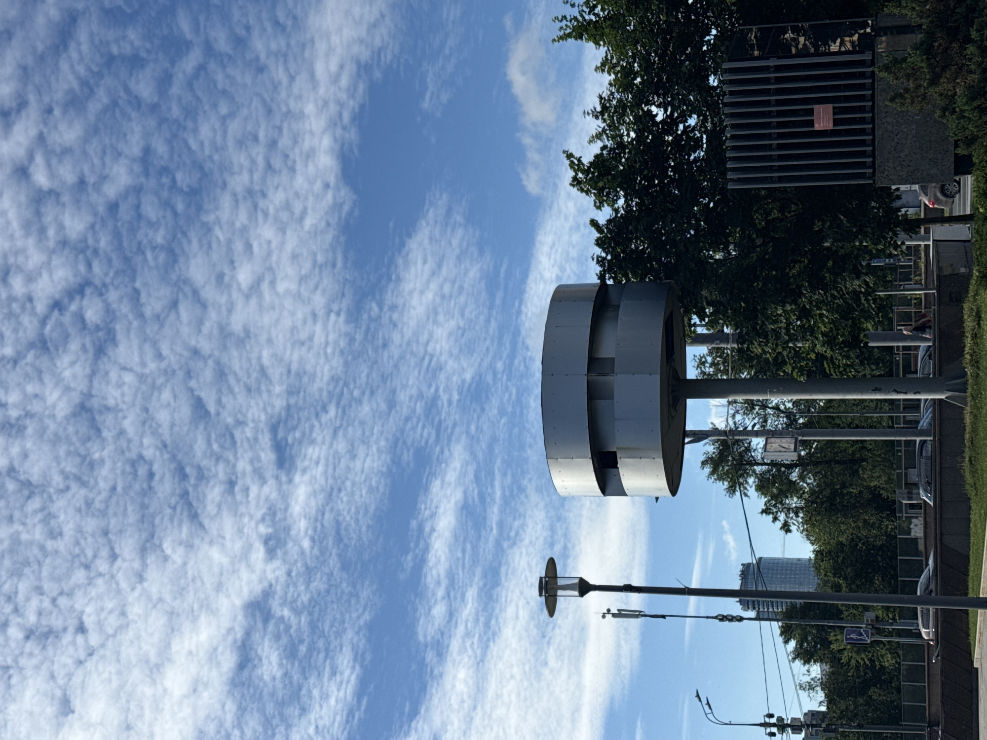 A cylindrical, metallic structure with two horizontal openings stands on a pole in an urban environment. A street lamp is also visible, as are trees, buildings, and a sky filled with white, wispy clouds. Cars are present on a road below. The scene is located in Moscow, Russia.