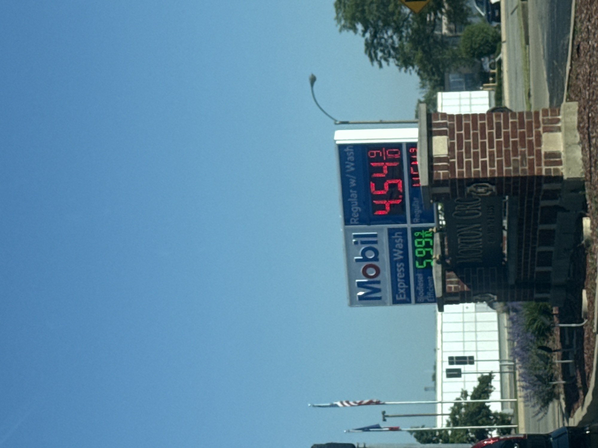 The image shows a Mobil gas station in Morton Grove, United States of America. A sign displays the prices for regular gas at $4.54 per gallon and express wash biodiesel efficient fuel at $5.99 per gallon. The sign also indicates "Regular with wash" at the top. The Morton Grove sign can be seen to the right of the Mobil gas sign. Behind the sign, there is a white building with large windows. In the background, there are trees and a clear blue sky. American flags and other poles can be seen further below the sign. A street light curves above the sign.