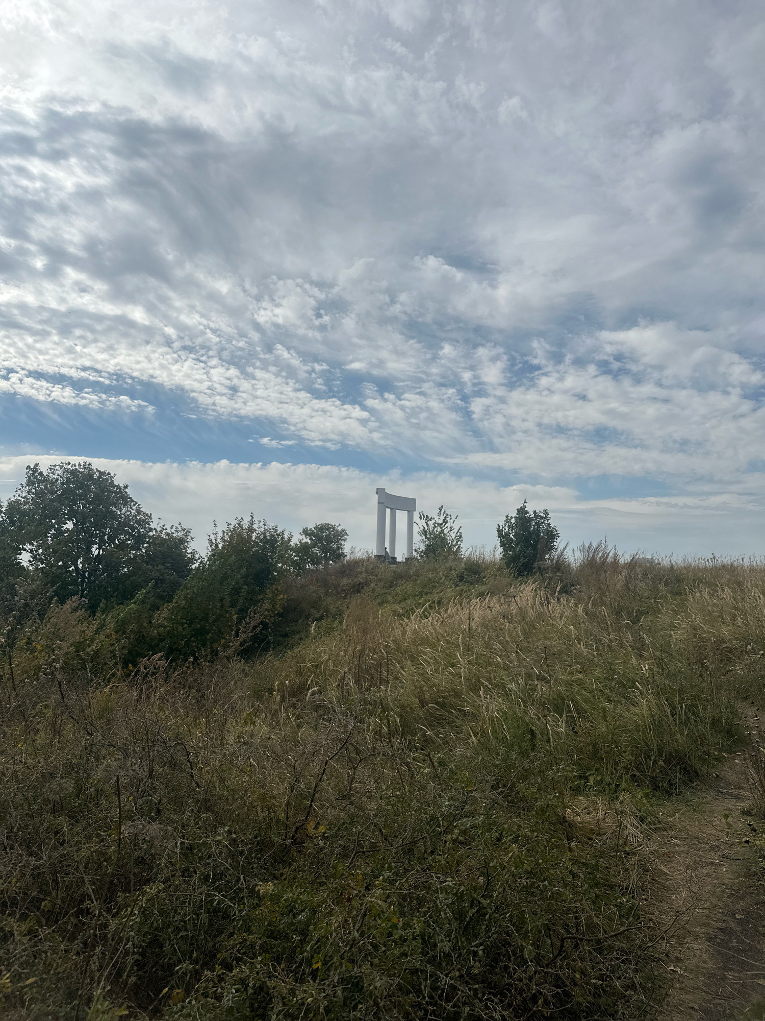 An exterior, eye-level, wide shot captures a landscape with a monumental structure on a grassy hill under a cloudy sky. The sky is filled with wispy, white clouds against a light blue background. In the middle ground, a white, abstract architectural monument composed of three columns supporting a horizontal beam stands on the crest of the hill. The monument is surrounded by scattered trees and bushes. The foreground is dominated by tall, dry grass and low-lying vegetation, with a dirt path winding through it on the right. The overall impression is one of a rural or park-like setting. The location is identified as Simskii, Russia. There are no people or animals clearly visible in the frame.