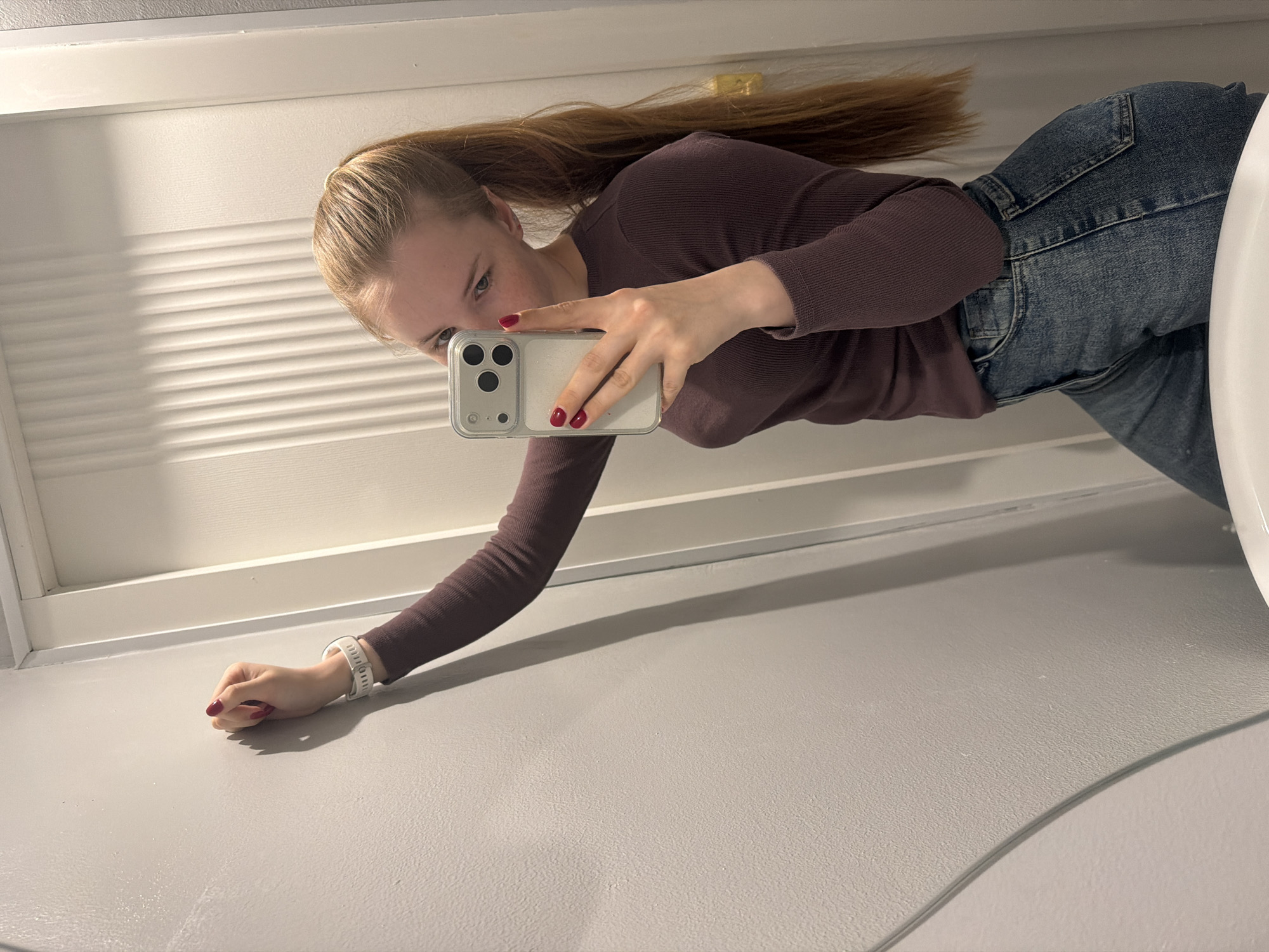A young woman with blonde hair pulled back into a ponytail is taking a selfie in a bathroom. She is wearing a maroon long-sleeved shirt and blue jeans. She is holding a smartphone with a clear case in her right hand to take the picture. Her nails are painted a dark red. Her left hand is resting on a gray countertop or vanity surface, and she's wearing a white wristband. The background consists of a white-framed mirror, a gray surface, and what appears to be a portion of a white sink. No information is given to determine if the location is in the city of Berezniki, Russia.