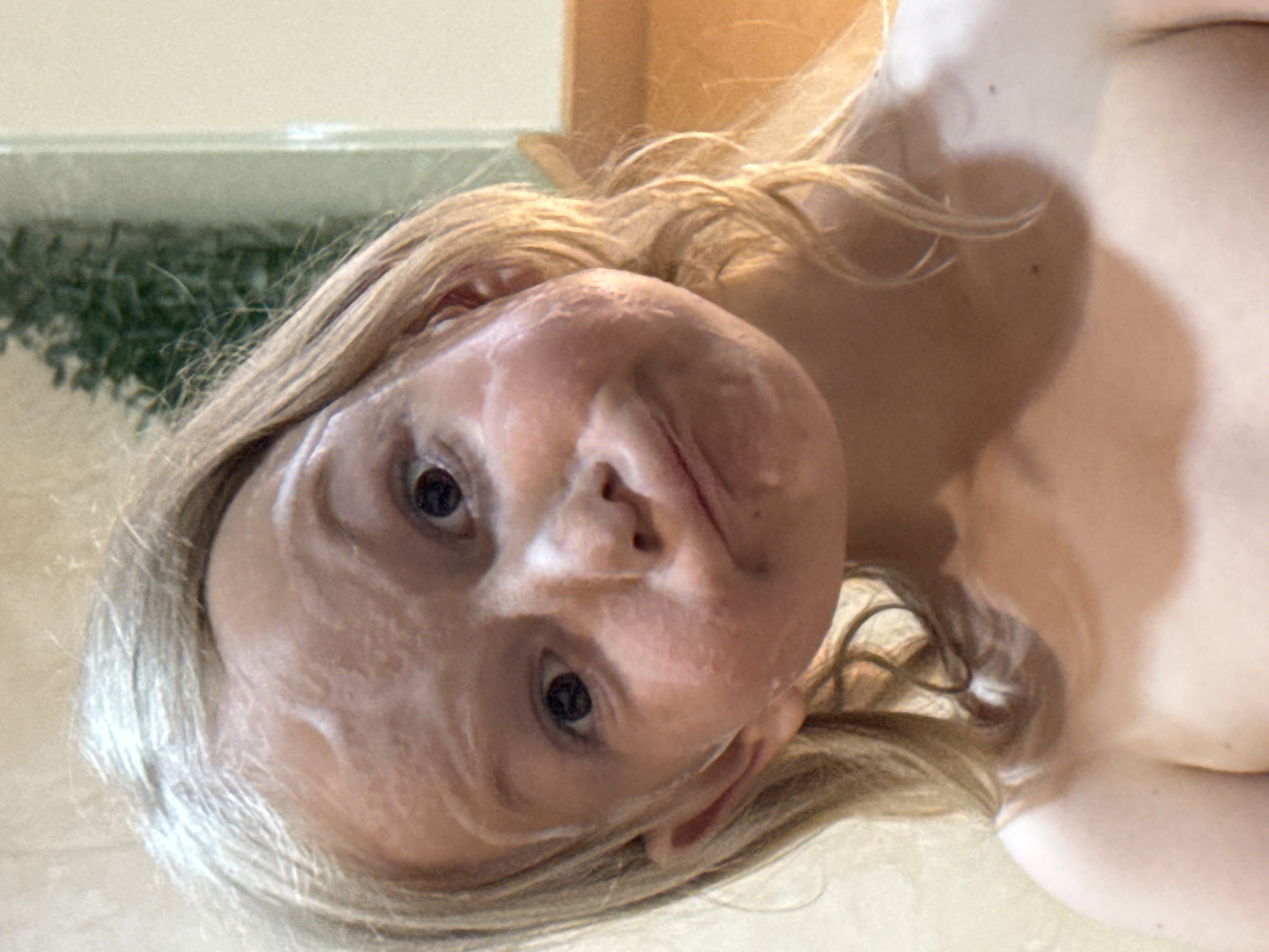A close-up, slightly angled view captures a person with fair skin and blonde hair, positioned against a neutral background. The face appears to have a thick white cream applied. The person is indoors and location is Unknown, Unknown.