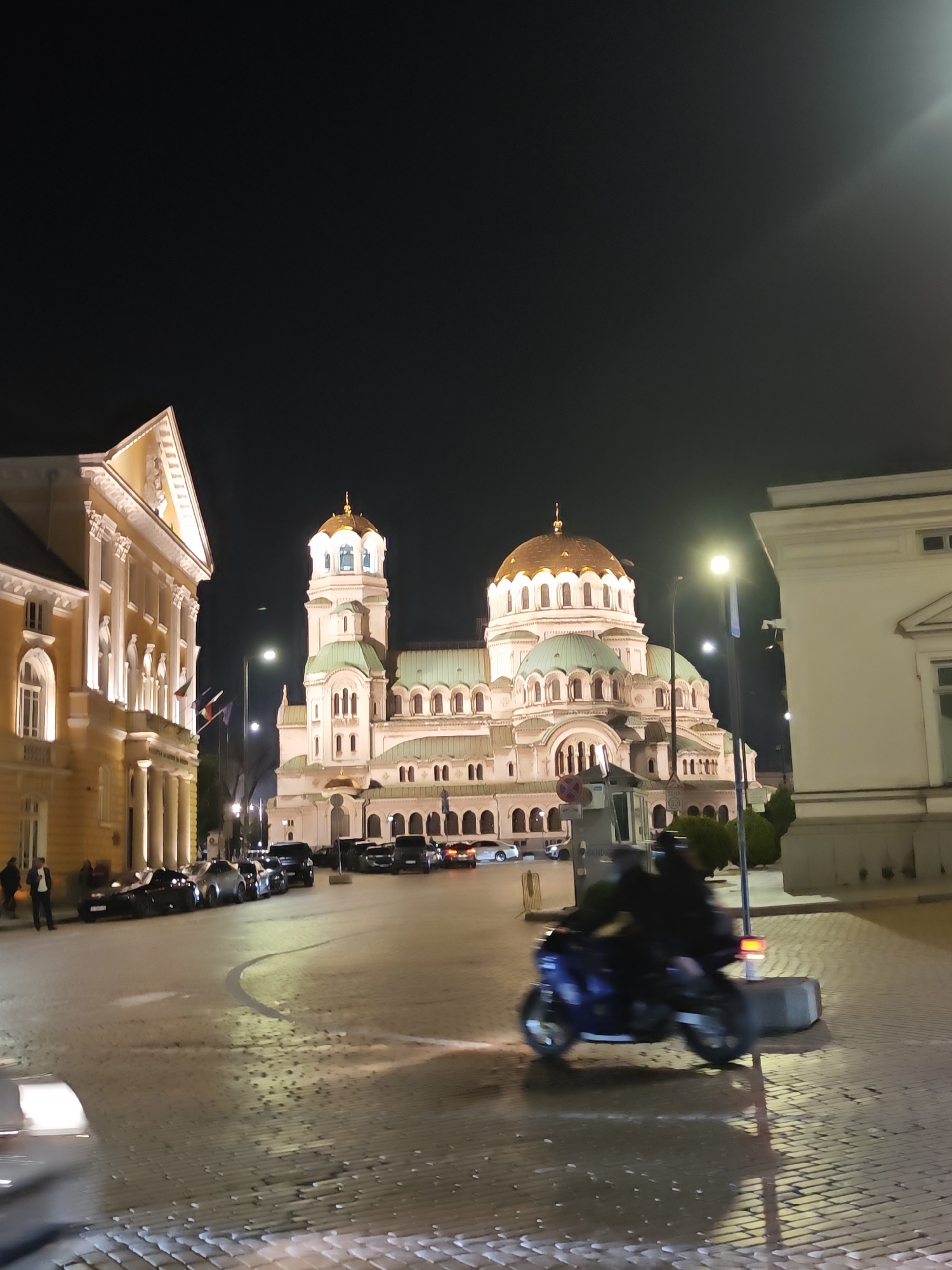 The image shows a nighttime scene in Sofia, Bulgaria, featuring the Alexander Nevsky Cathedral illuminated in the background. To the left is a yellow building with a columned facade. Parked cars line the street between the two buildings and the cathedral. In the foreground, a motorcycle is blurred in motion, indicating its speed. The street is paved with cobblestones. Streetlights illuminate the area, and people are visible in front of the yellow building to the left. The sky is dark, and the architectural details of the buildings are highlighted by the artificial lighting.