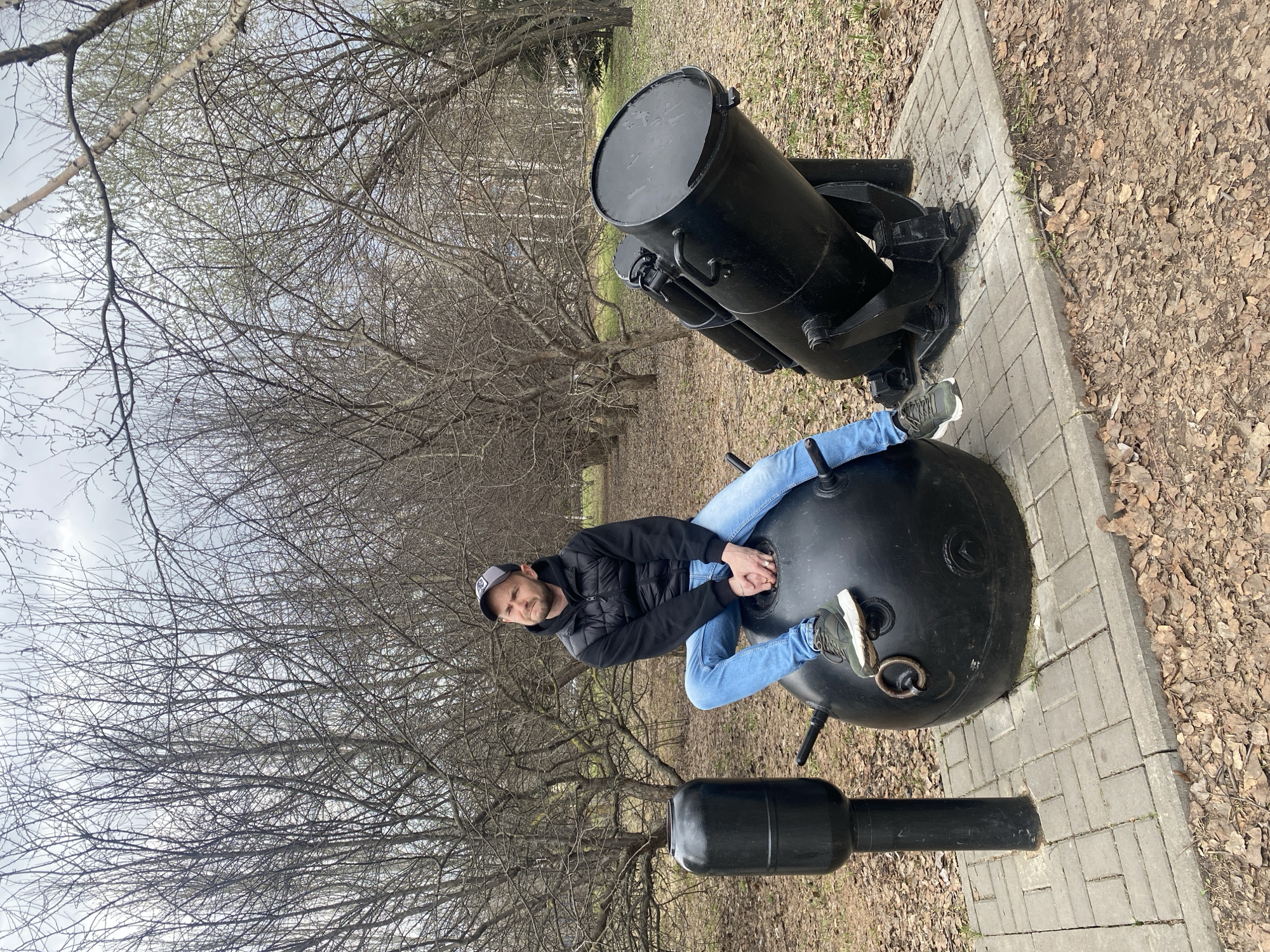 A man in a black jacket and blue jeans sits atop a black spherical object with protrusions in an outdoor setting. His legs are bent, and he appears relaxed. Behind him, there is a large, black cylindrical object mounted on a black frame, resting on a paved surface made of rectangular bricks. The ground is covered in dry leaves, and bare trees are visible in the background under a cloudy sky. The location appears to be in the city of Kazan, Russia, as indicated by the naval mine and mortar, which are part of a memorial complex dedicated to naval warfare.