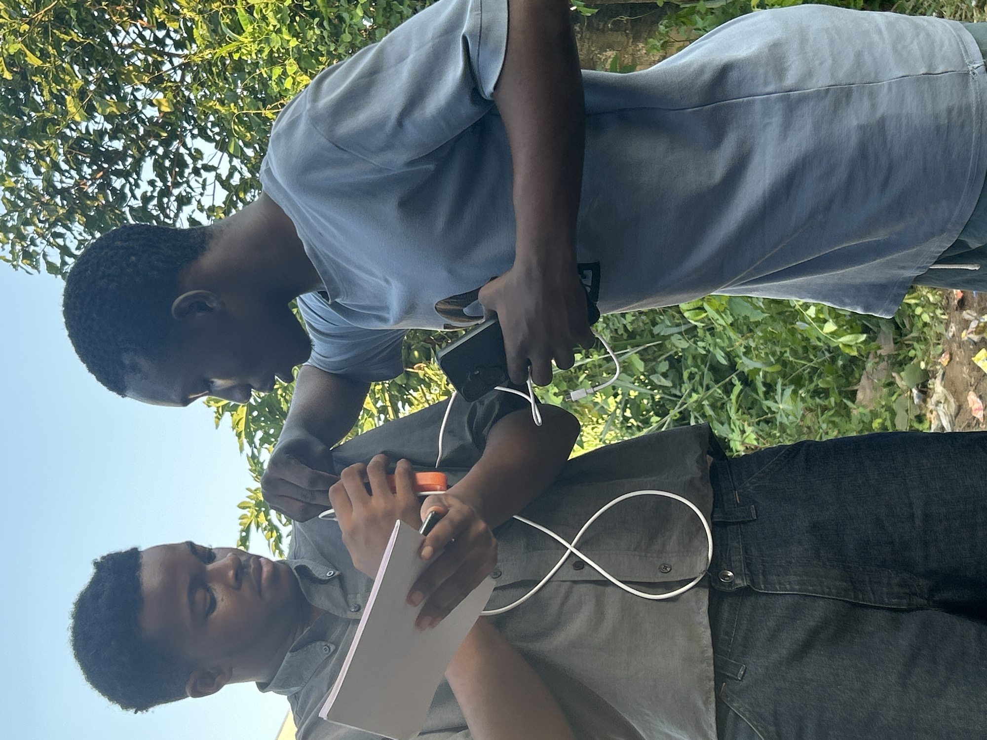 Two young Black males are standing outdoors amidst lush green foliage and a clear sky. The male on the left, wearing a dark collared shirt and dark denim jeans, is looking down and holding a white notepad and a pen. A white charging cable, plugged into an orange portable charger, is looped around his neck and connected to a device out of frame. The male on the right, wearing a light blue t-shirt and dark shorts, is looking towards the other male and holding a black smartphone in his right hand, with a white charging cable connected to it. The scene appears to be a casual outdoor setting, possibly in a residential area of Ihiagwa, Nigeria.