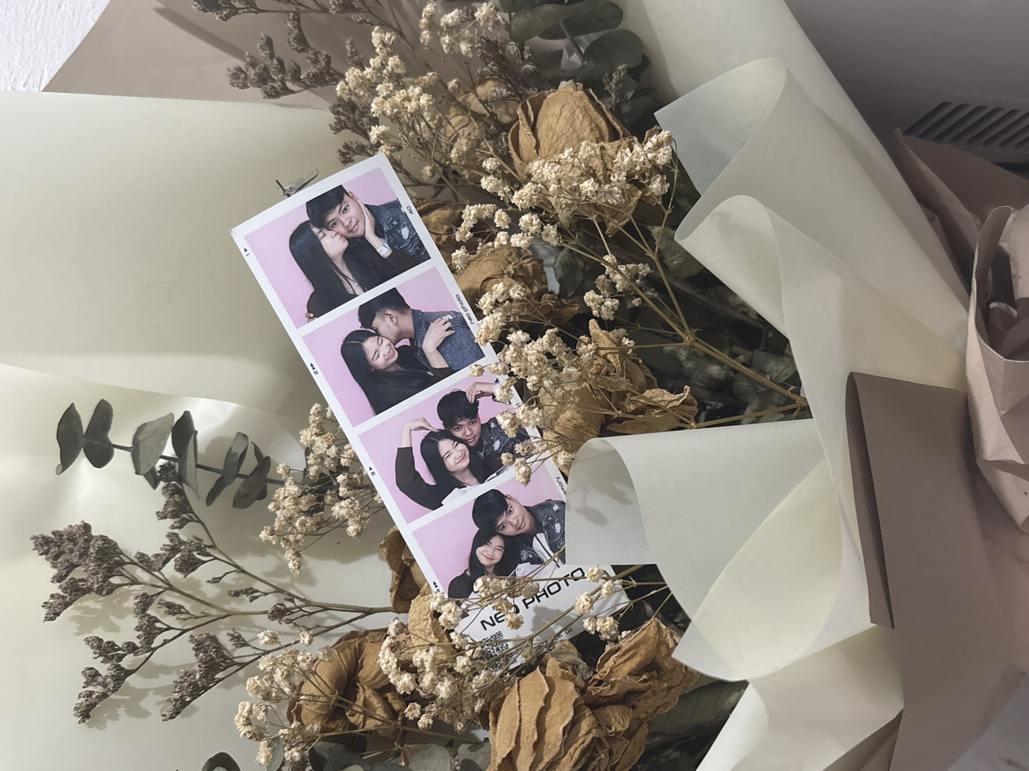 A bouquet of dried flowers is arranged in a light-colored wrapping paper, with a strip of photo booth photos featuring a young Asian couple inserted into the bouquet. The photos show the couple in various poses, including kissing and hugging. The couple appears to be in their late teens or early twenties. The background is a light-colored wall, suggesting an indoor setting. The image does not contain any specific landmarks or details to pinpoint a location within North Jakarta, Indonesia.