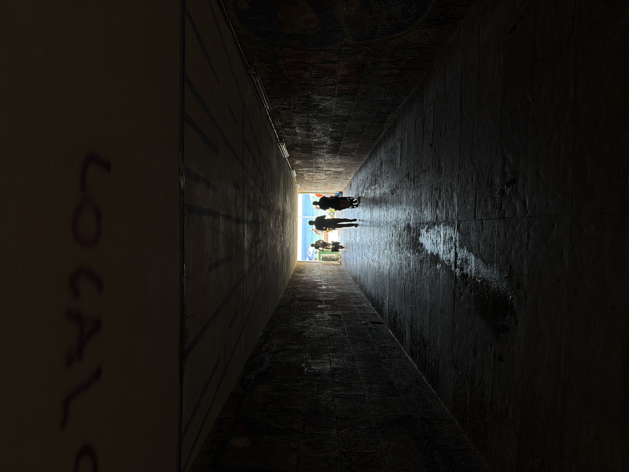 A view of a dark tunnel in Cascais, Portugal, leading to an opening with bright, blue light. Silhouettes of a few individuals are visible near the exit. The walls of the tunnel are made of dark stone or brick, with graffiti on the left wall reading "LOCAL." The tunnel floor appears to be slightly wet.