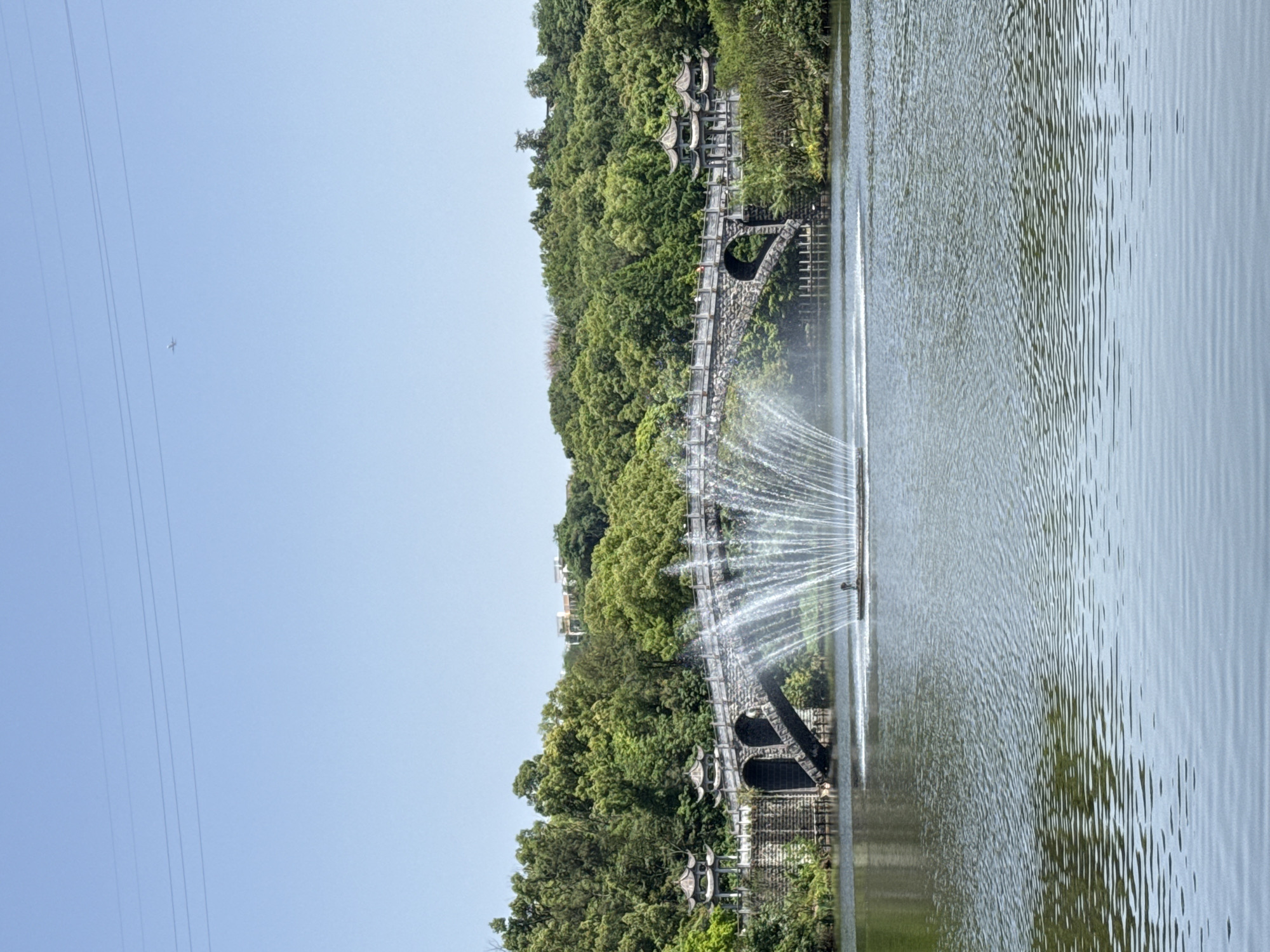 The image captures a serene scene at the Lake Scenic Area of Xijiao Park in Jiulongpo District, Chongqing, China. A bridge with arched openings, partially obscured by lush green trees, stands to the left of a body of water. In the water, a fountain is active, spraying water upward. The sky is clear blue, with a faint trace of an airplane visible in the upper left and electrical wires in the same vicinity. The water exhibits ripples, and the overall lighting suggests a bright, possibly sunny, day.