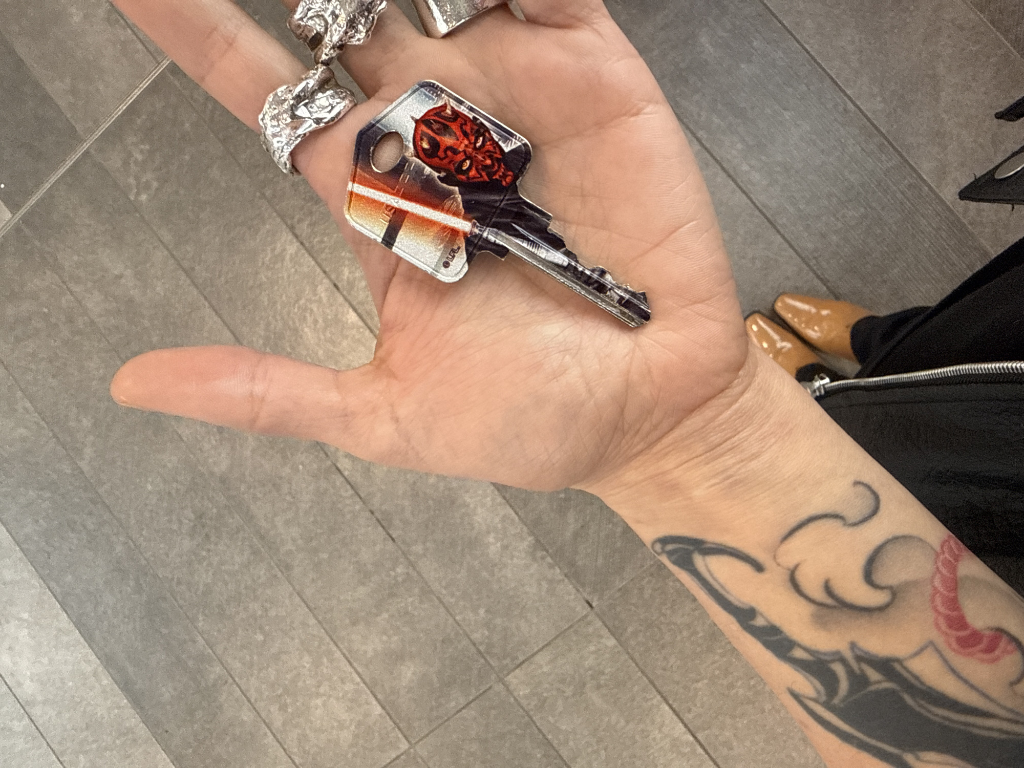 A person holds a key with a Star Wars image (Darth Maul) in their left palm. The person has rings on their fingers and a tattoo on their forearm. The setting appears to be indoors with tiled flooring. In the background, a glimpse of the person's tan shoes and black clothing is visible. The image was taken in District of North Vancouver, Canada.