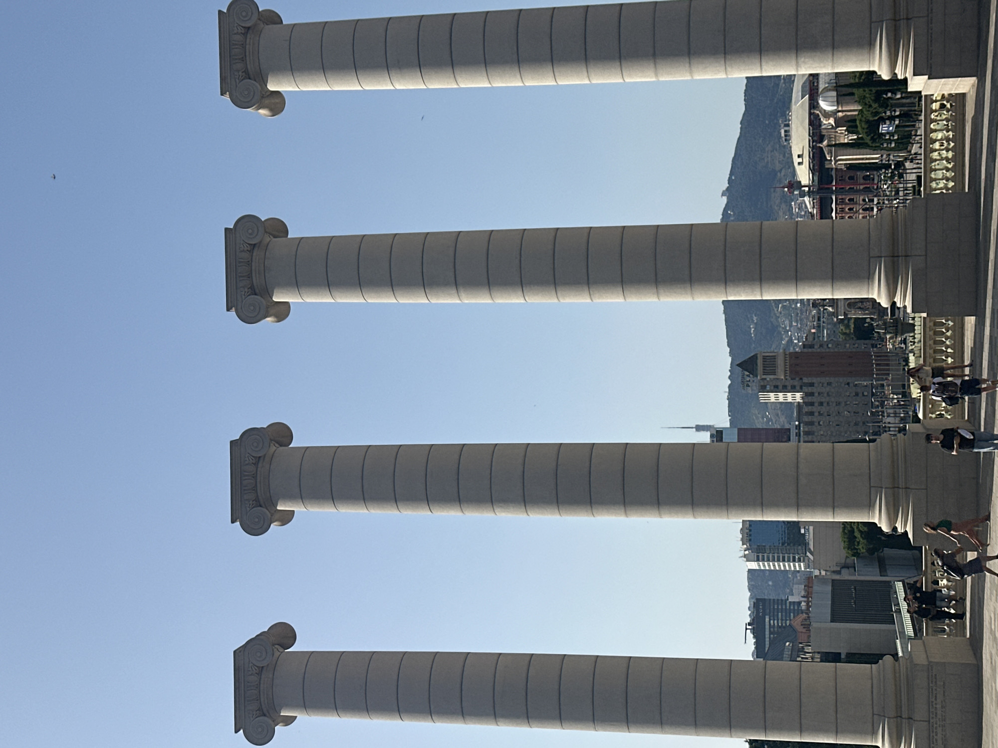 The image shows a view of the Four Columns of Puig i Cadafalch, located at the foot of Montjuïc hill in Barcelona, Spain. The four Corinthian columns, constructed from granite, are imposing and symmetrical, with fluted shafts and ornate capitals. They stand in the foreground, framing a panoramic view of the city.
In the background, a cityscape unfolds under a clear blue sky. Various buildings of different architectural styles and heights are visible, including modern skyscrapers and older structures. A prominent tower with a spire can be seen, along with a large, domed building that resembles an arena or stadium. Mountains rise in the distance, forming a backdrop to the urban landscape.
At the base of the columns, a paved area leads to a balustrade, and beyond that, the city stretches out. A few people are present in the scene, standing on the paved area and near the balustrade. Some are casually dressed, and their interactions appear relaxed. The overall impression is one of a bright, sunny day in a vibrant urban setting, with classical architecture providing a striking contrast to the modern city.