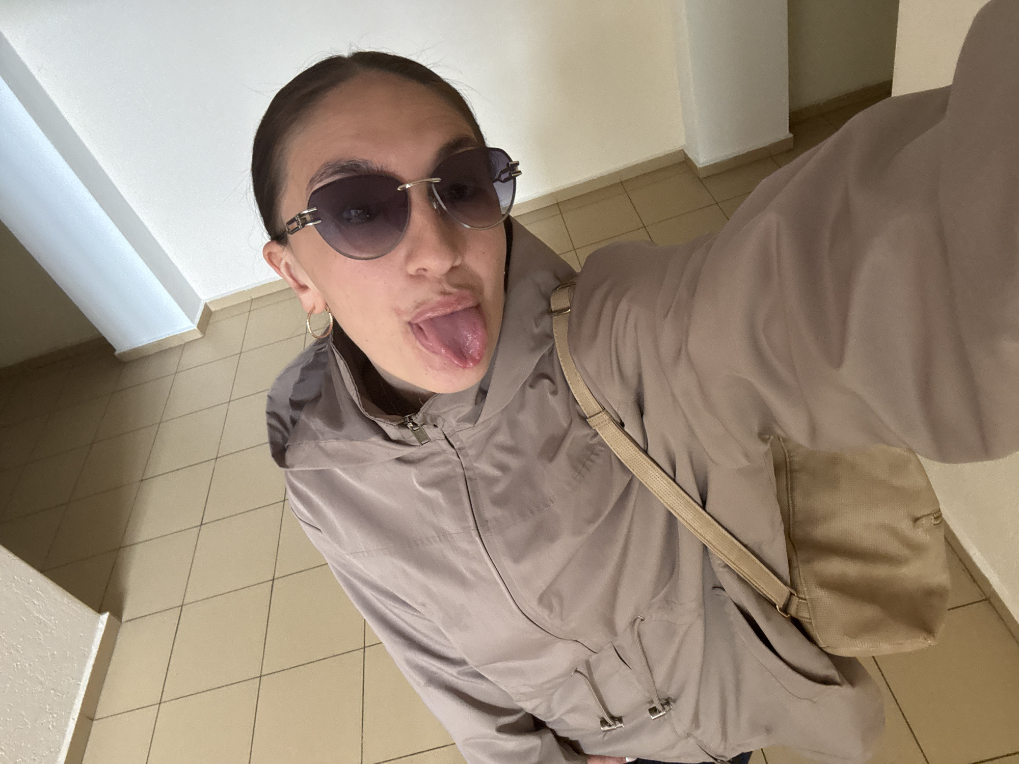 A woman wearing sunglasses and a beige jacket is sticking out her tongue while taking a selfie in an interior setting. She has a purse slung over her shoulder and is standing on tiled flooring. The location appears to be a hallway or lobby with white walls. The specific city location of Sochi, Russia cannot be determined from the image alone.