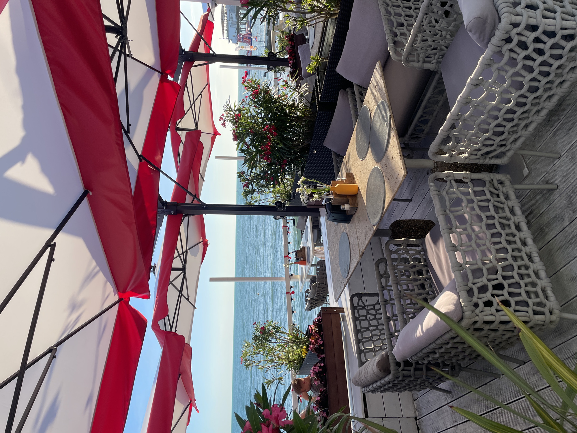 This image depicts an outdoor seating area at a seaside restaurant or cafe, likely in Sochi, Russia. The scene is bathed in the warm, golden light of late afternoon or early evening. Large red and white striped umbrellas provide shade over several tables and chairs. The furniture appears to be modern and made of grey wicker with light purple cushions. On one table, placemats and a yellow bottle are visible.
In the background, the calm, turquoise sea stretches to the horizon, under a clear sky. A few boats or watercraft can be seen in the distance. Lush green plants with vibrant red and pink flowers are placed in planters, adding color and life to the setting. A person is partially visible in the lower right quadrant of the image, seemingly interacting with the plants. The overall atmosphere suggests a relaxing and enjoyable dining experience by the sea. There is no visible text in the image.