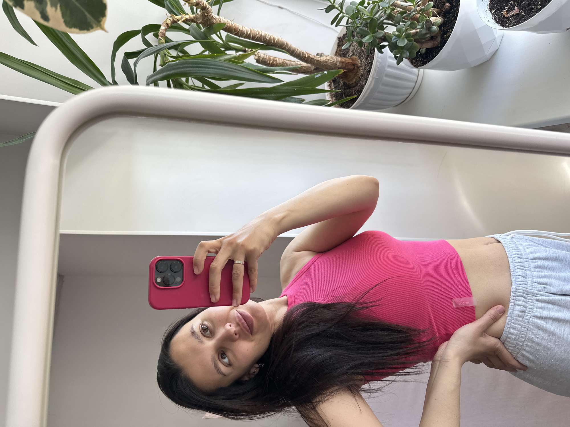 A woman is taking a selfie in a mirror. She is wearing a pink tank top and gray sweatpants. She is holding a pink phone in her right hand. There are several plants in white pots visible above the mirror. The background appears to be a white wall. There is no evidence to indicate that the image was taken in Chaikovsky, Russia.