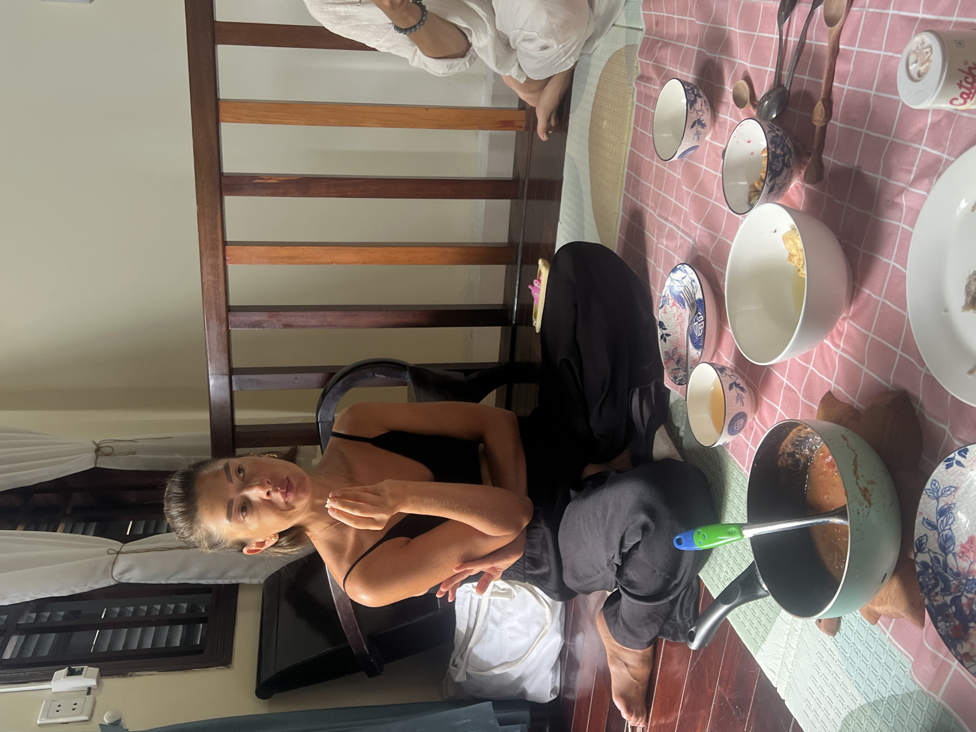 In Da Nang, Vietnam, a woman with short brown hair and a black tank top and pants sits cross-legged on a pink checkered mat laid on a dark wooden floor. She is holding a small white object between her thumb and forefinger, appearing to be in the process of eating or examining it. Her right arm is bent, with her hand near her mouth. The woman is seated in a room with dark wooden furniture, including a chair behind her and a railing in the background.
On the mat in front of her are several bowls, some containing food items. One larger, light-colored bowl contains a yellow liquid and some white pieces. Another smaller bowl has a blue and white pattern and contains small, light-colored nuts or seeds. A turquoise pot with a green ladle, containing a reddish sauce, is positioned to the left. Other bowls and wooden spoons are scattered around the mat.
To the right, a person wearing light-colored clothing, possibly white linen, is also seated on the mat, though their face is not visible. The background shows a window with white curtains and dark blinds, and a light-colored wall. The overall scene suggests a casual meal or gathering in a home setting.