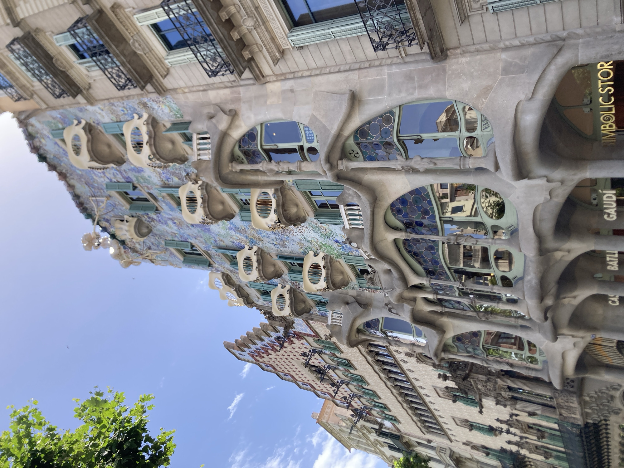 This is a photograph of Casa Batlló, a famous building designed by Antoni Gaudí, located in Barcelona, Spain. The image is taken from a low angle, looking up at the building's facade. The architecture is characterized by its organic, flowing lines and colorful mosaic details.
The facade features unique windows with bone-like structures surrounding them, and a roof adorned with mosaic tiles that resemble scales or dragon skin. The colors are vibrant, with blues, greens, and purples dominating the mosaic work. Several windows show reflections of the surrounding buildings and the sky. Visible text on the building includes "GAUDI" and "SYMBOLIC STORE".
The sky is blue with some scattered white clouds, indicating a clear day. The sun appears to be shining, casting light on the building and creating reflections. The presence of lush green trees in the foreground suggests a pleasant, possibly warm, weather condition, ideal for outdoor sightseeing. The photograph captures the distinctive and whimsical style of Gaudí's architecture, making it a striking visual representation of this landmark.