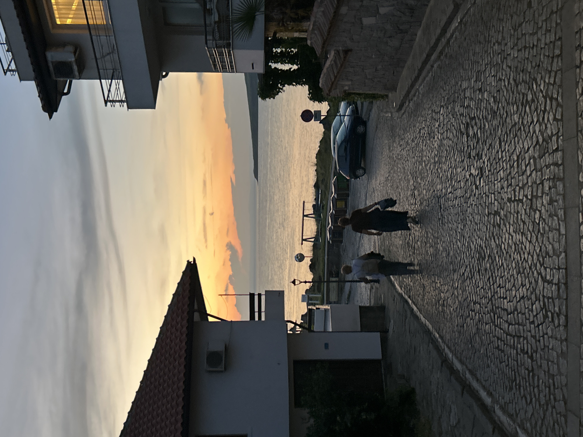 The image depicts a cobblestone street in Sozopol, Bulgaria, leading towards the sea at dusk. Two individuals are walking away from the viewer down the street. The person in front is wearing a dark dress and carrying a bag, while the person behind them is wearing a light-colored shirt and has a backpack. On the left side of the street is a white building with a red-tiled roof. On the right, another building, also white with balconies, is visible, along with a stone wall. The sea stretches out in the background, with a distant landmass visible on the horizon. The sky is filled with the warm colors of sunset, with clouds adding texture to the scene. Several cars are parked along the right side of the street. A street lamp and some playground equipment can be seen closer to the sea. The overall impression is of a peaceful evening in a coastal town.