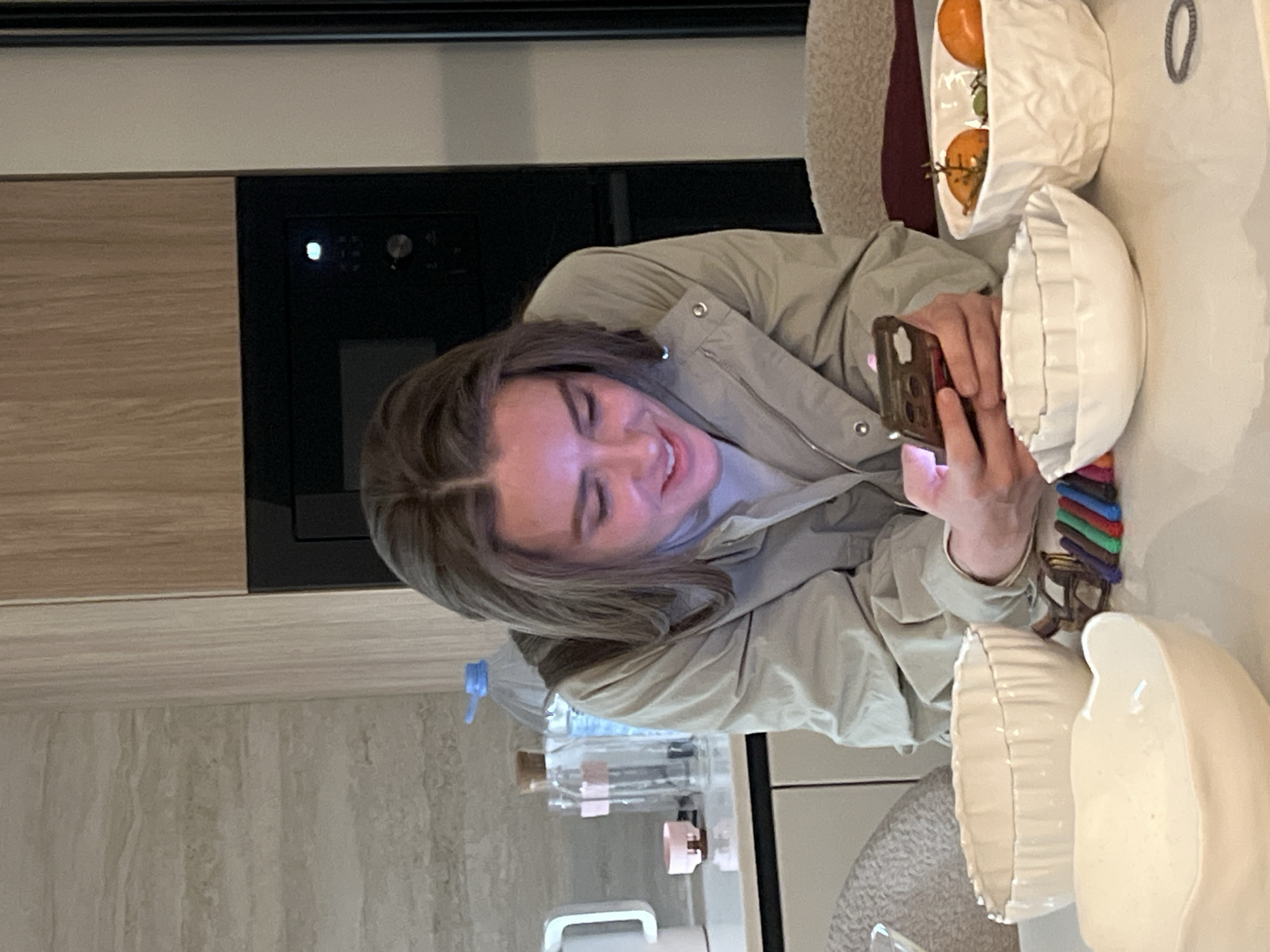 A woman in a light tan jacket is seated at a table in an indoor environment. She is looking at and holding a smartphone. On the table are three white decorative bowls, one containing oranges. Multiple water bottles are visible near the woman. The location is not determinable as Рубцово, Russia with the given information.