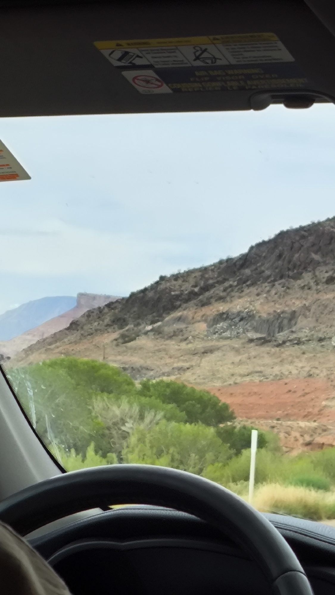 The image is taken from inside a vehicle looking out the windshield at a desert landscape. A steering wheel is visible in the foreground. Beyond the windshield, there are rolling hills covered in dry brush and scattered green vegetation. In the distance, a larger, dark-colored mountain range dominates the horizon. The sky is overcast with a pale blue hue. A white signpost stands in the middle ground to the right. The location is identified as Virgin, United States of America.